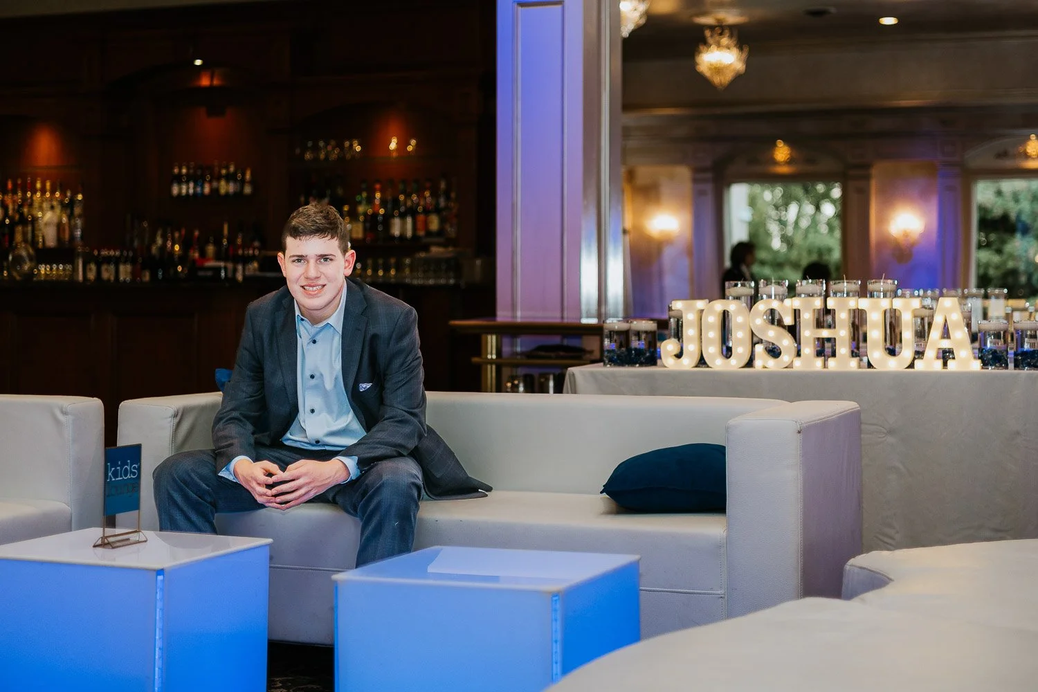 A young man in a suit sitting on a white sofa at a decorated indoor event space, with a lit display spelling 'JOSHUA' in the background and a bar area to the left.