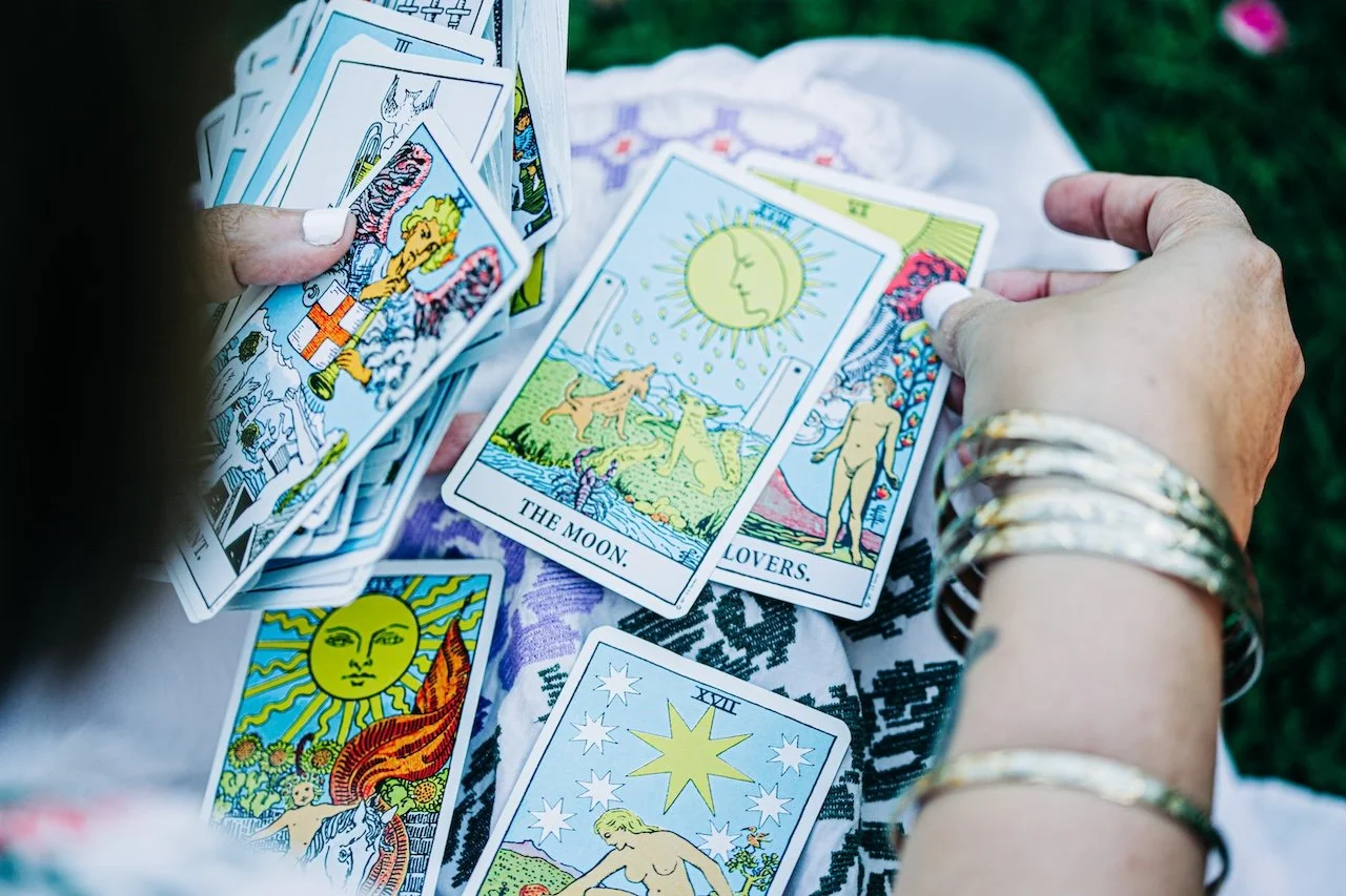 Person holding tarot cards outdoors, with several tarot cards laid out on a cloth on the ground. The cards feature colorful illustrations, including a card with the moon and two lovers.