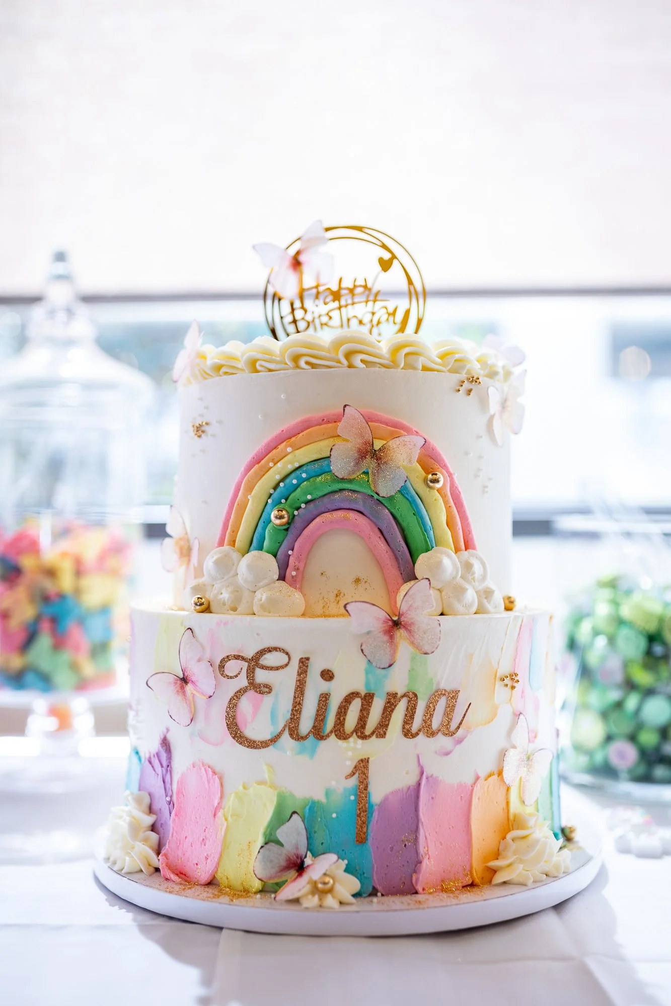 A colorful birthday cake with a rainbow design, decorated with pink butterflies, white roses, and a "Happy Birthday" topper. The cake has the name "Eliana" and the age "1" written on it.