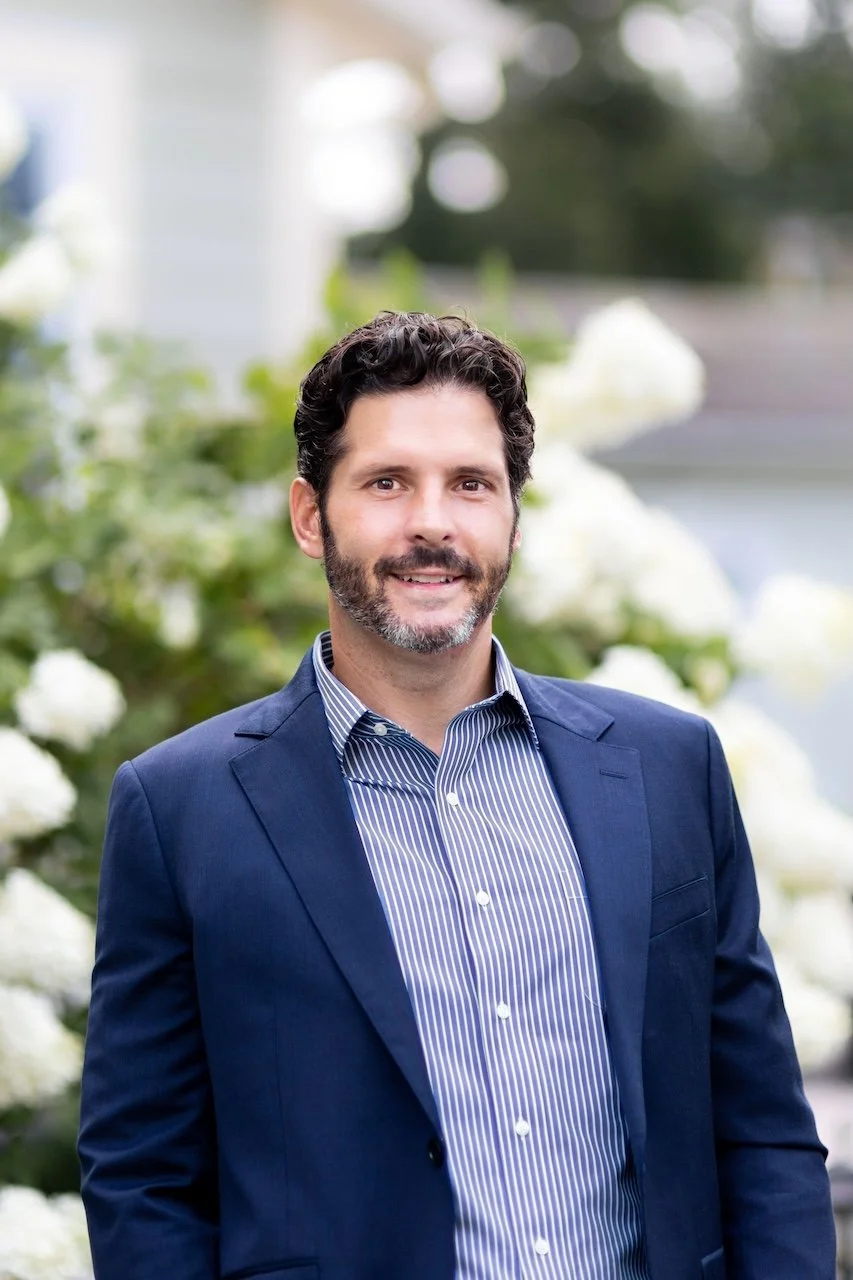 A man with dark, curly hair and a beard, wearing a navy suit and a blue-and-white striped shirt, standing outdoors with white flowers and greenery in the background.