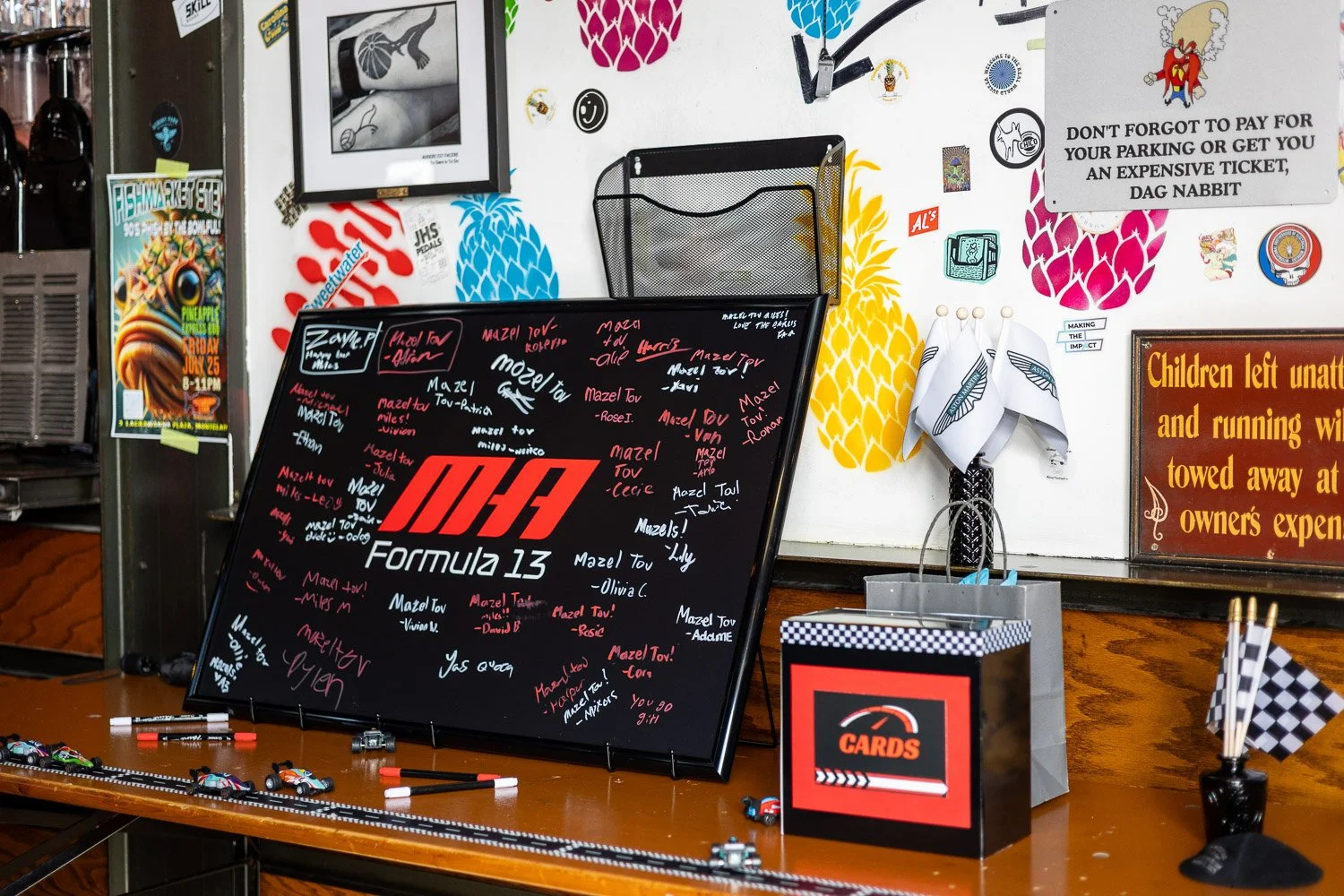 A racing-themed decor with a black blackboard featuring colorful signatures and a bold red and white logo, model cars on the table, and flags. Stickers and signs on the wall, including a motivational sign and a parking reminder. A small gift bag labe