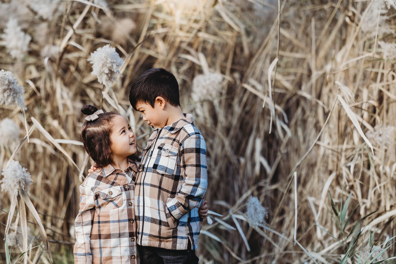 A young girl and boy, both wearing plaid shirts, stand close together in a field of tall, dry grasses and fluffy plants, smiling and looking at each other.
