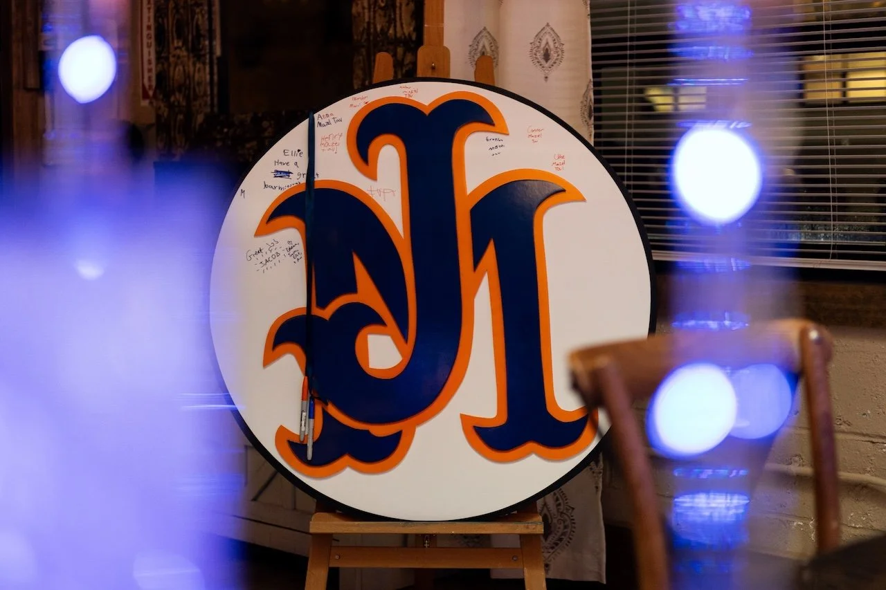 Round sign with a large blue and orange stylized letter 'L' at the center, covered in handwritten messages, placed on an easel in front of a window with blinds, with some blue and white bokeh lights in the foreground.