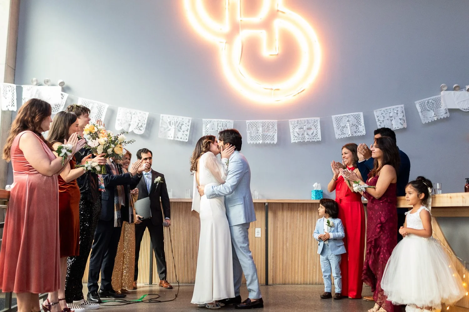 A bride and groom kiss during their wedding ceremony in front of friends and family at a brewery in Brooklyn, with a neon Power button sign above and decorative banners on the wall.