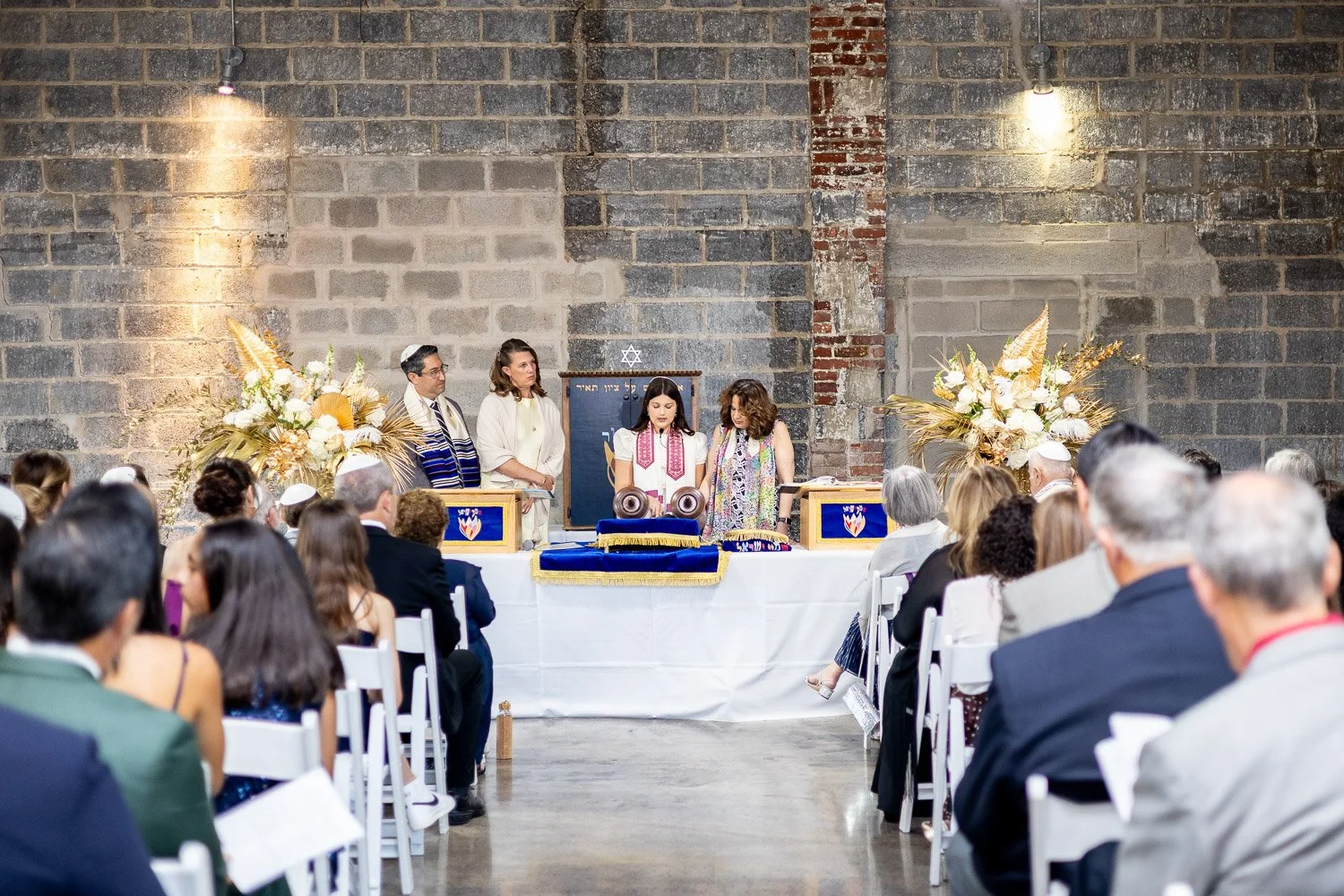 Jewish wedding ceremony with rabbis and rabbis and guests seated in front of a brick wall, with floral arrangements and a Torah scroll on a table.