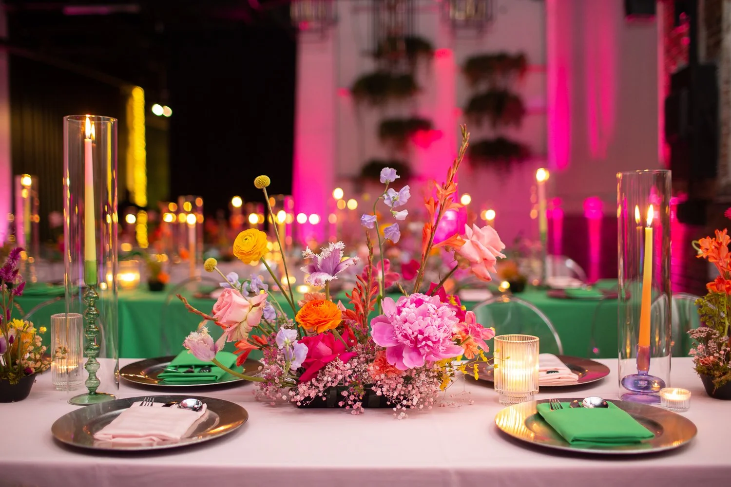 Decorated event table with colorful flowers, tall candles in glass holders, and green napkins, with glowing pink and yellow lighting in the background.