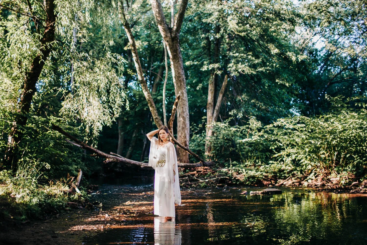 A woman wearing a long white dress and earrings standing in a shallow creek surrounded by lush green trees in a forest.