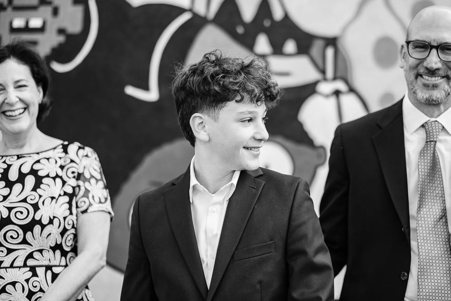 Three people, two men and one woman, standing in a row and smiling, with a colorful mural in the background.