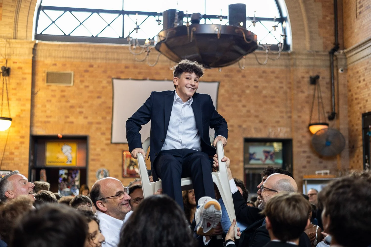 A young man in a suit sitting on a chair being lifted above a crowd in an indoor event space with exposed brick walls.