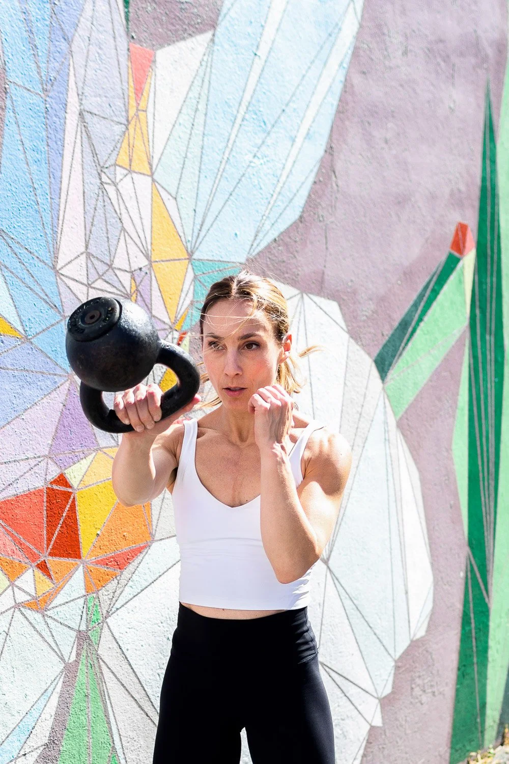A woman in workout attire, holding a kettlebell in an outdoor gym area with a colorful geometric mural on the wall behind her.