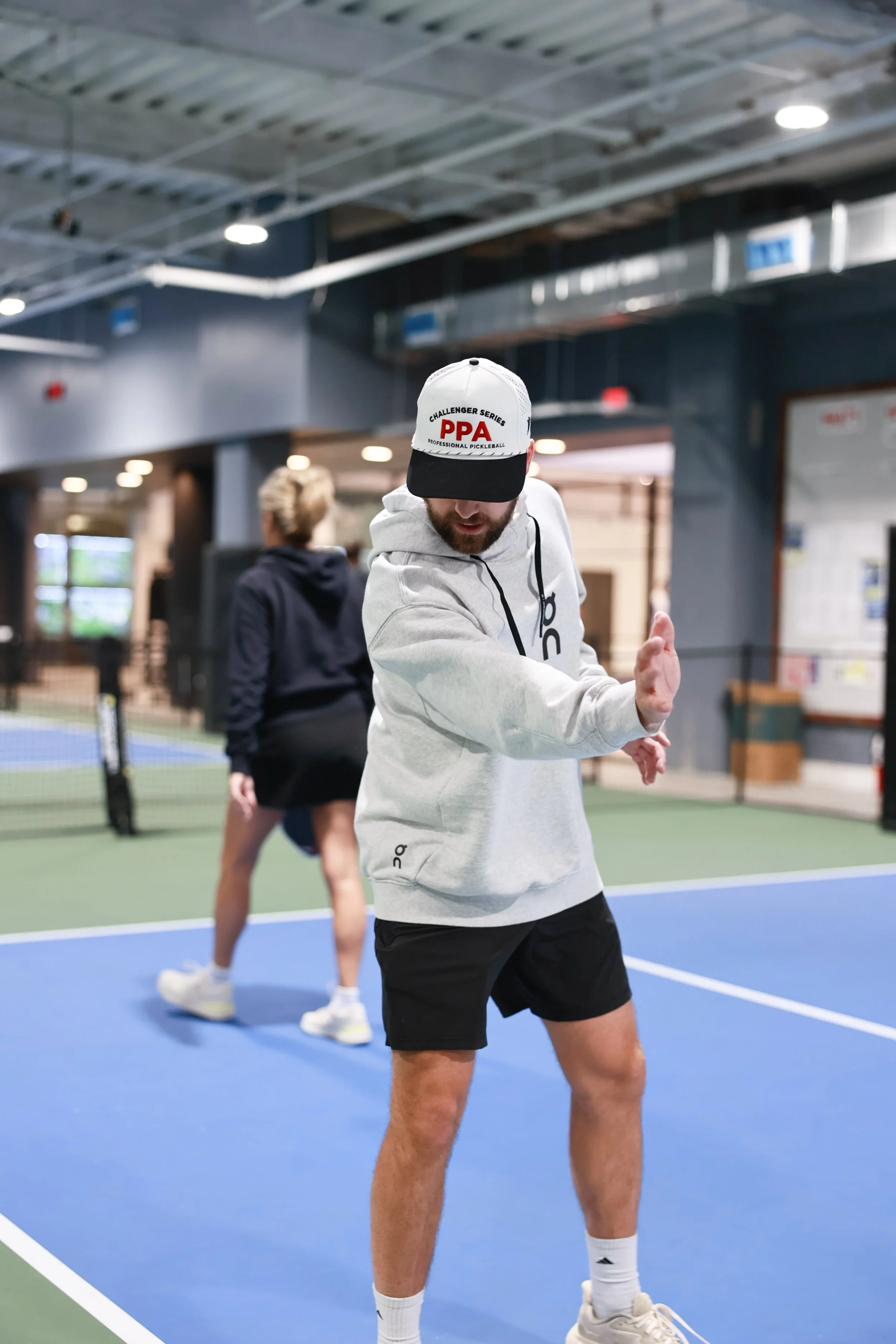 A man wearing a white hoodie, black shorts, and white sneakers is playing pickleball indoors. He is in a ready stance with his hand extended, and a woman in a black hoodie and shorts is in the background on the same court.