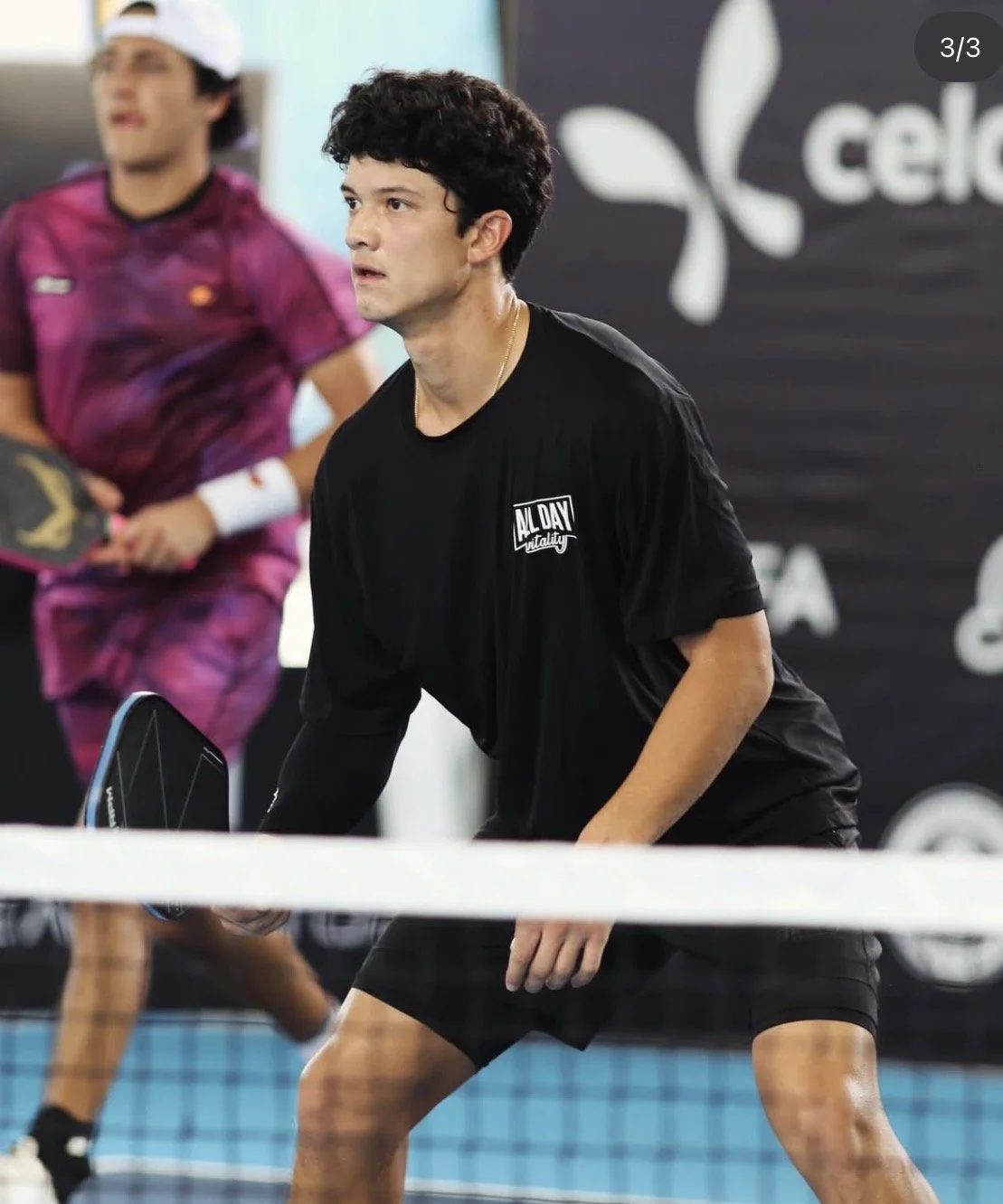 A young man playing pickleball, holding a paddle in a ready position, wearing a black T-shirt with 'All Day Vitality' printed on it, on an indoor court.