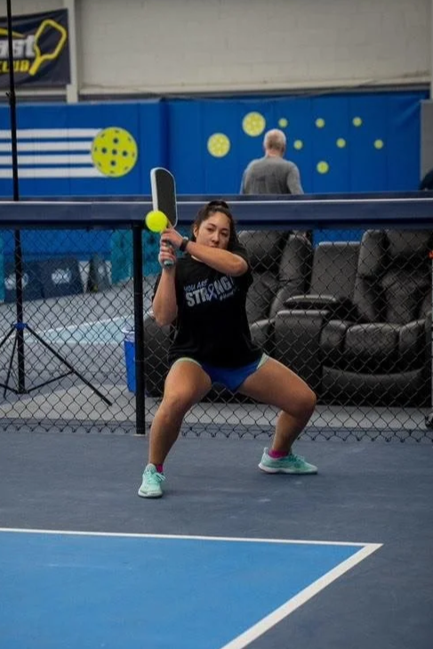 A woman practicing tennis moves on an indoor tennis court, holding a tennis racket and a tennis ball, with a net behind her.