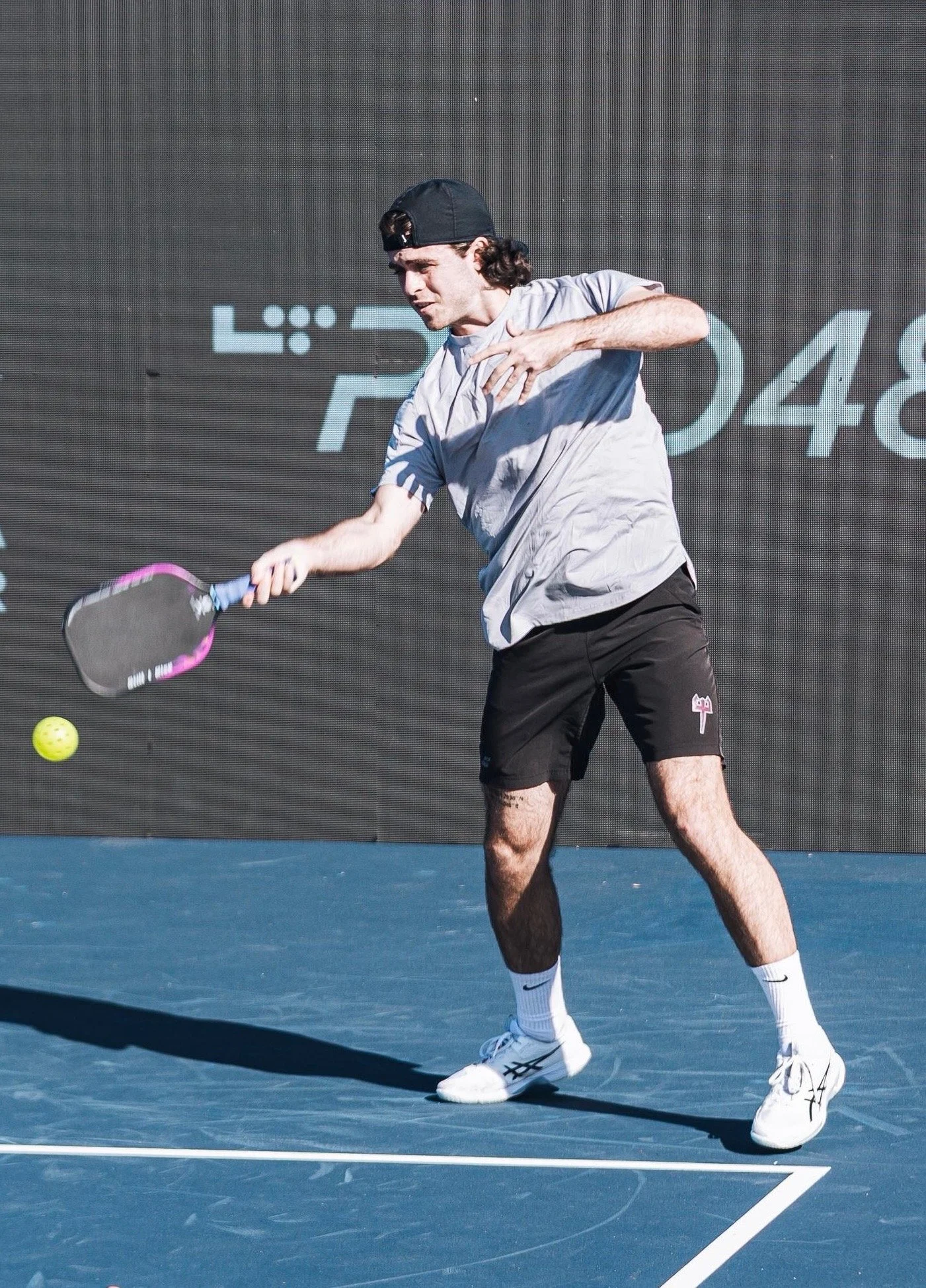 A man playing pickleball on a blue court, wearing a black cap, a gray t-shirt, black shorts, and white tennis shoes, as he prepares to hit a yellow pickleball with a pink-handled paddle.