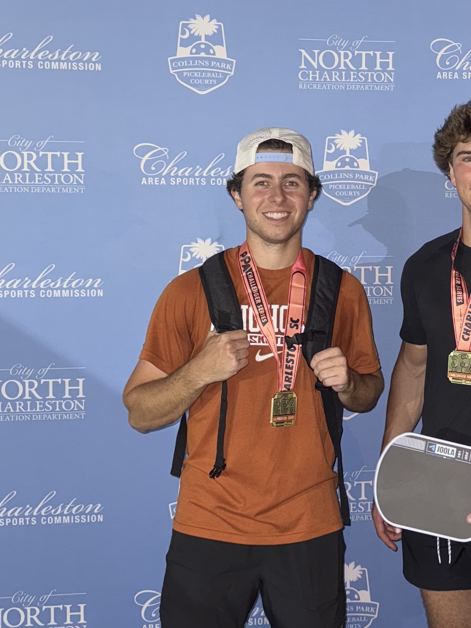Young man smiling, wearing a white cap backwards, an orange Nike t-shirt, carrying a black backpack, and wearing medals around his neck, standing in front of a blue backdrop with Charleston City logos.