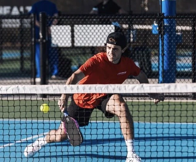 A young man in a red shirt and black shorts playing tennis on a blue court, preparing to strike a yellow tennis ball. He is wearing a black cap backwards, and there is a chain-link fence in the background.