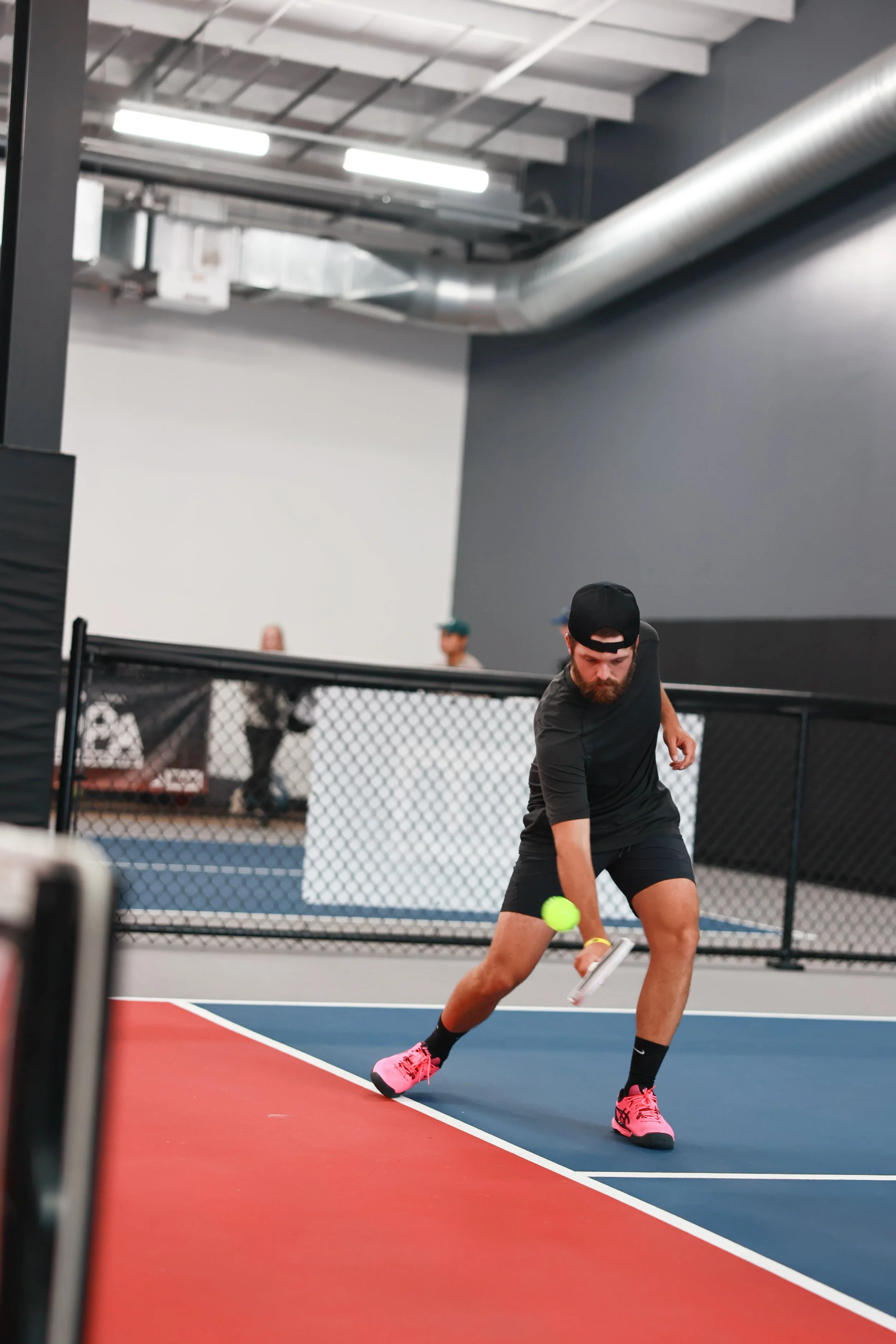 A man playing pickleball on an indoor court, dressed in black shorts, black shirt, pink shoes, and a black cap, preparing to hit a yellow pickleball with a paddle.