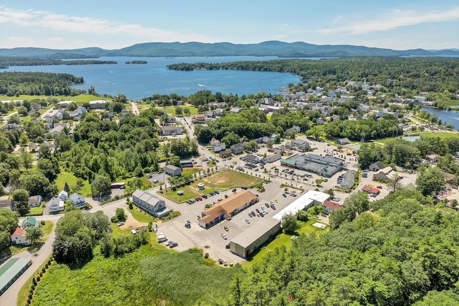 Aerial view of a small town near a large body of water, with green trees, houses, and parking lots on a sunny day.