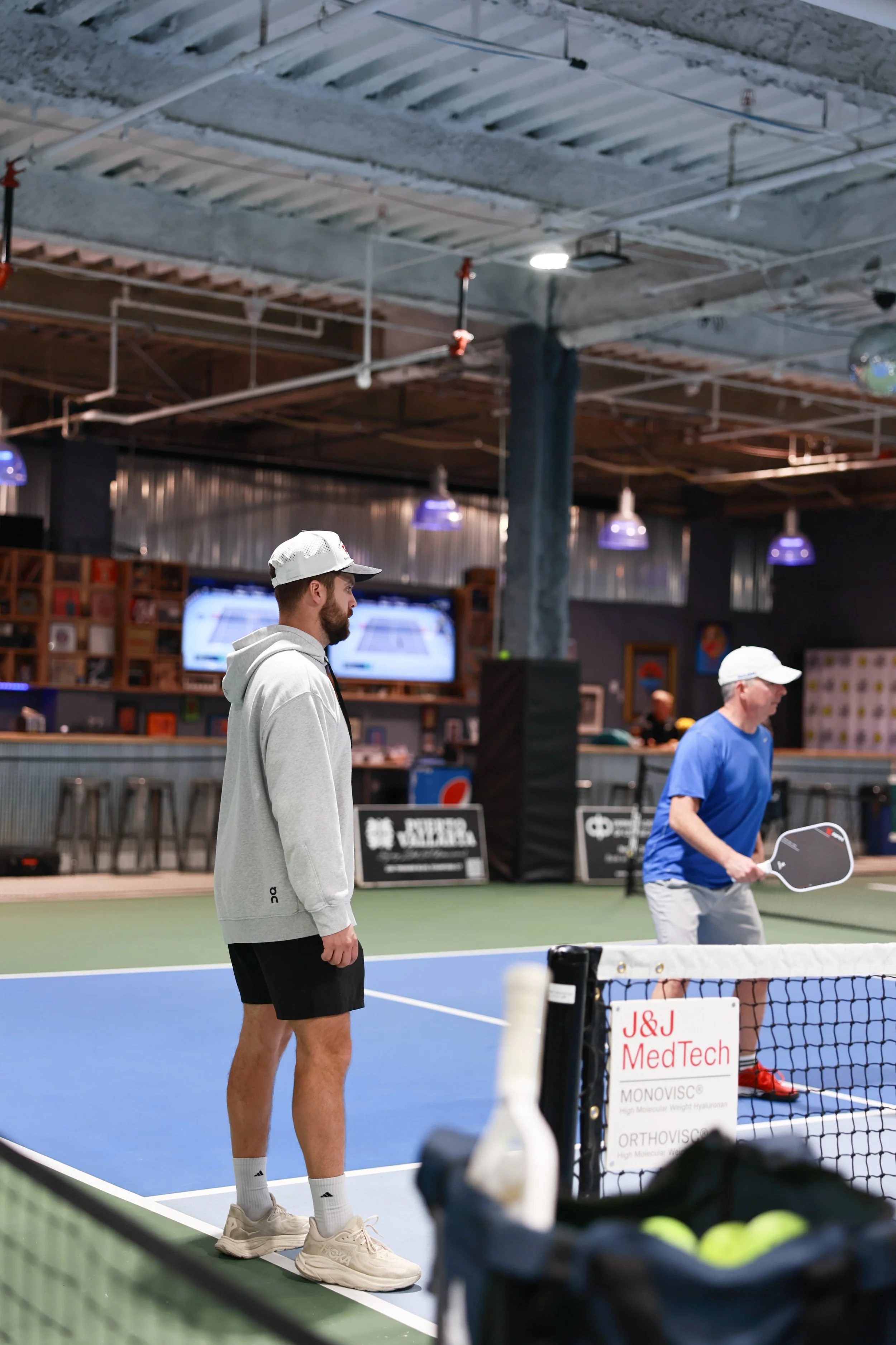 Two men playing pickleball on an indoor court, with one man preparing to hit the ball and the other watching, in a sports bar or lounge setting.
