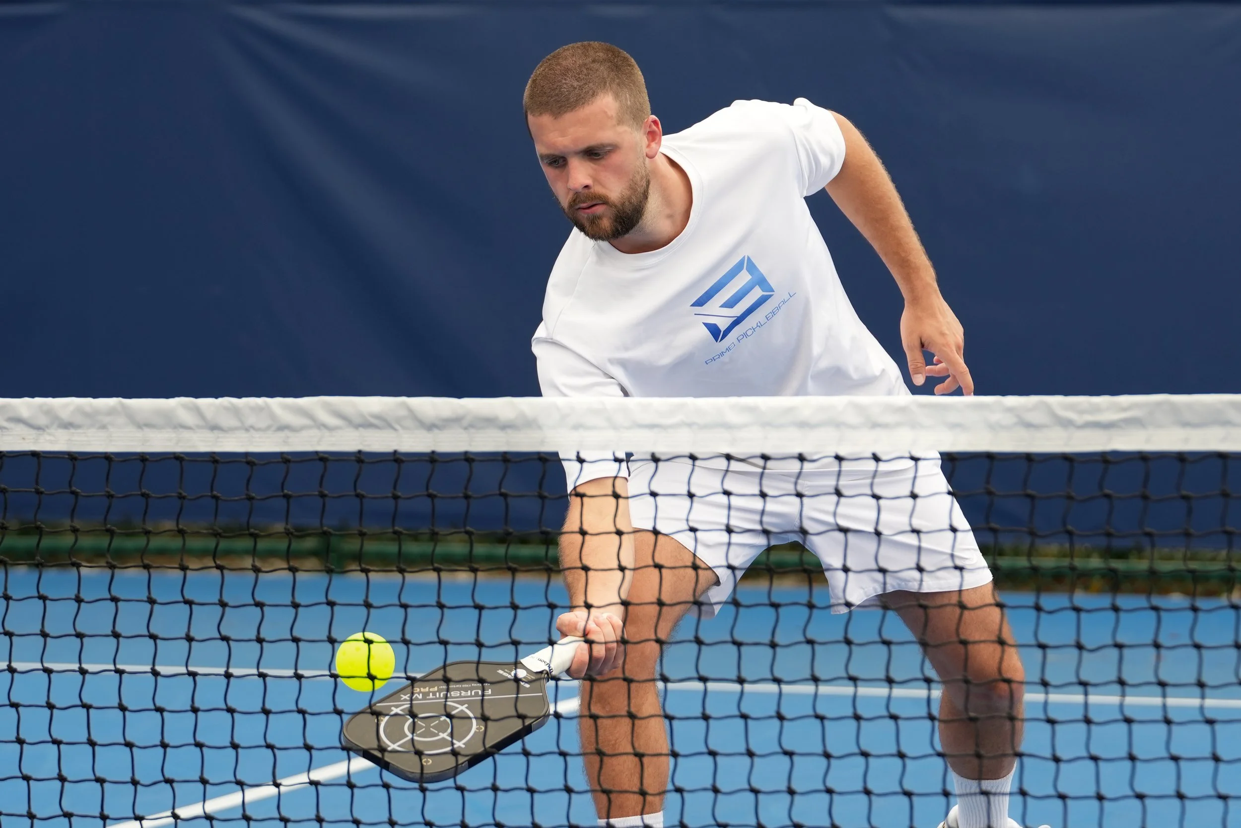 A man playing pickleball on an indoor court, hitting a yellow ball with a paddle while wearing a white T-shirt and white shorts.