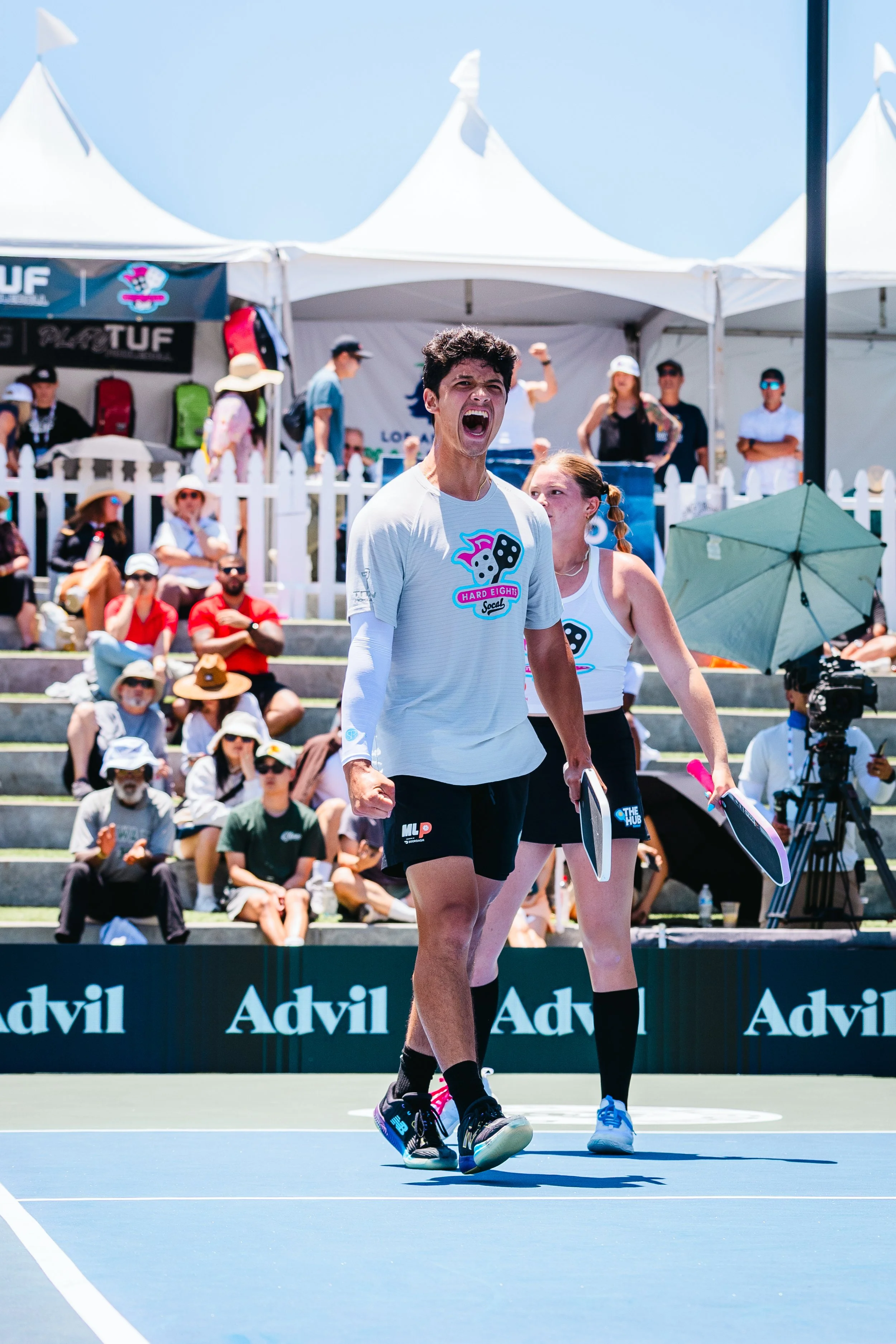 A young athlete celebrates on a tennis court during a match. He appears excited and victorious, holding a paddle in his right hand. Spectators sit in the background, some clapping and cheering, with tents and banners behind them.