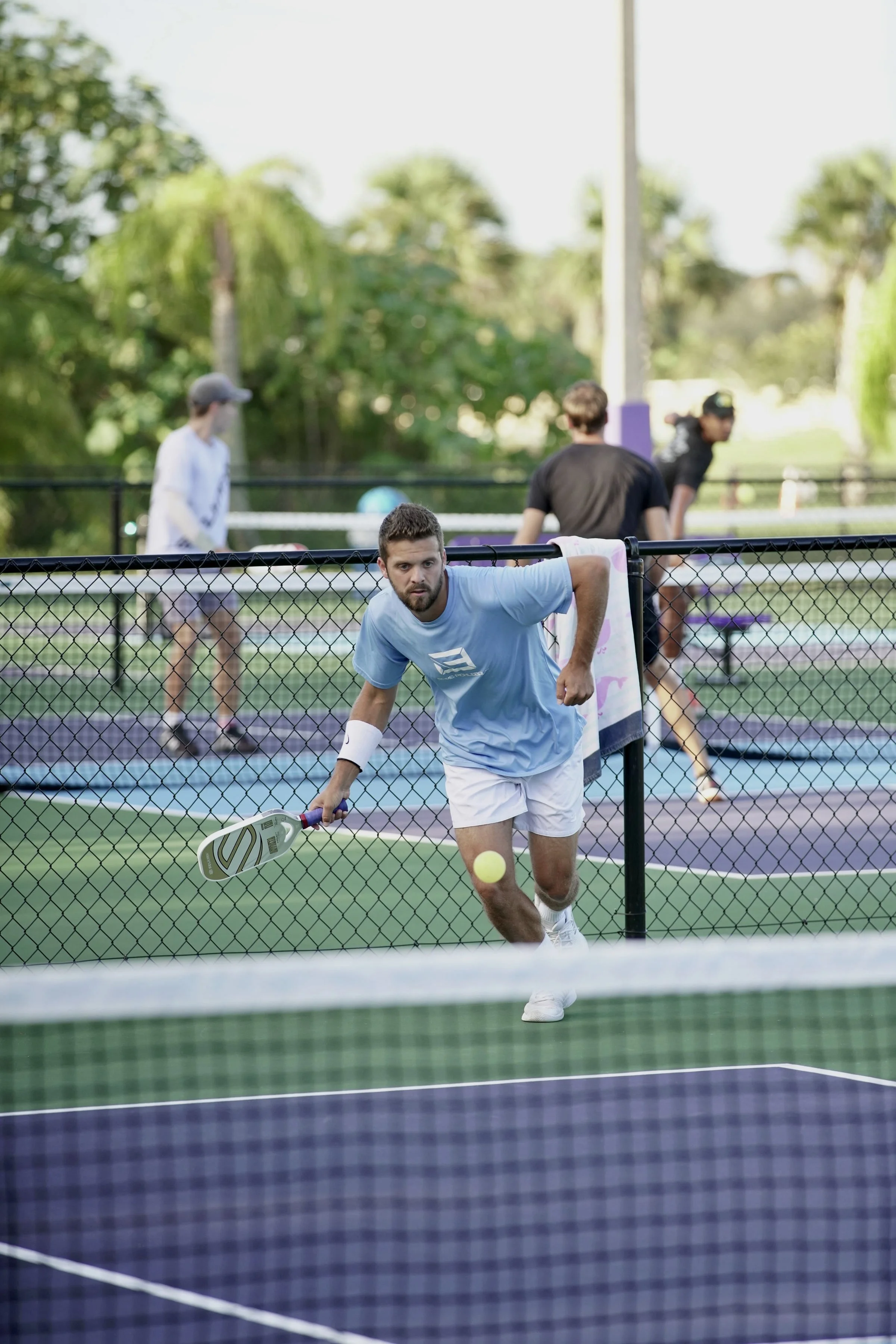 A man playing pickleball, hitting a ball with a paddle on an outdoor court, with other players in the background and trees surrounding.