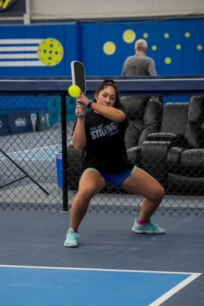A young woman is playing tennis on an indoor court, holding a tennis racket and preparing to hit a yellow tennis ball, with seating and a man in the background.
