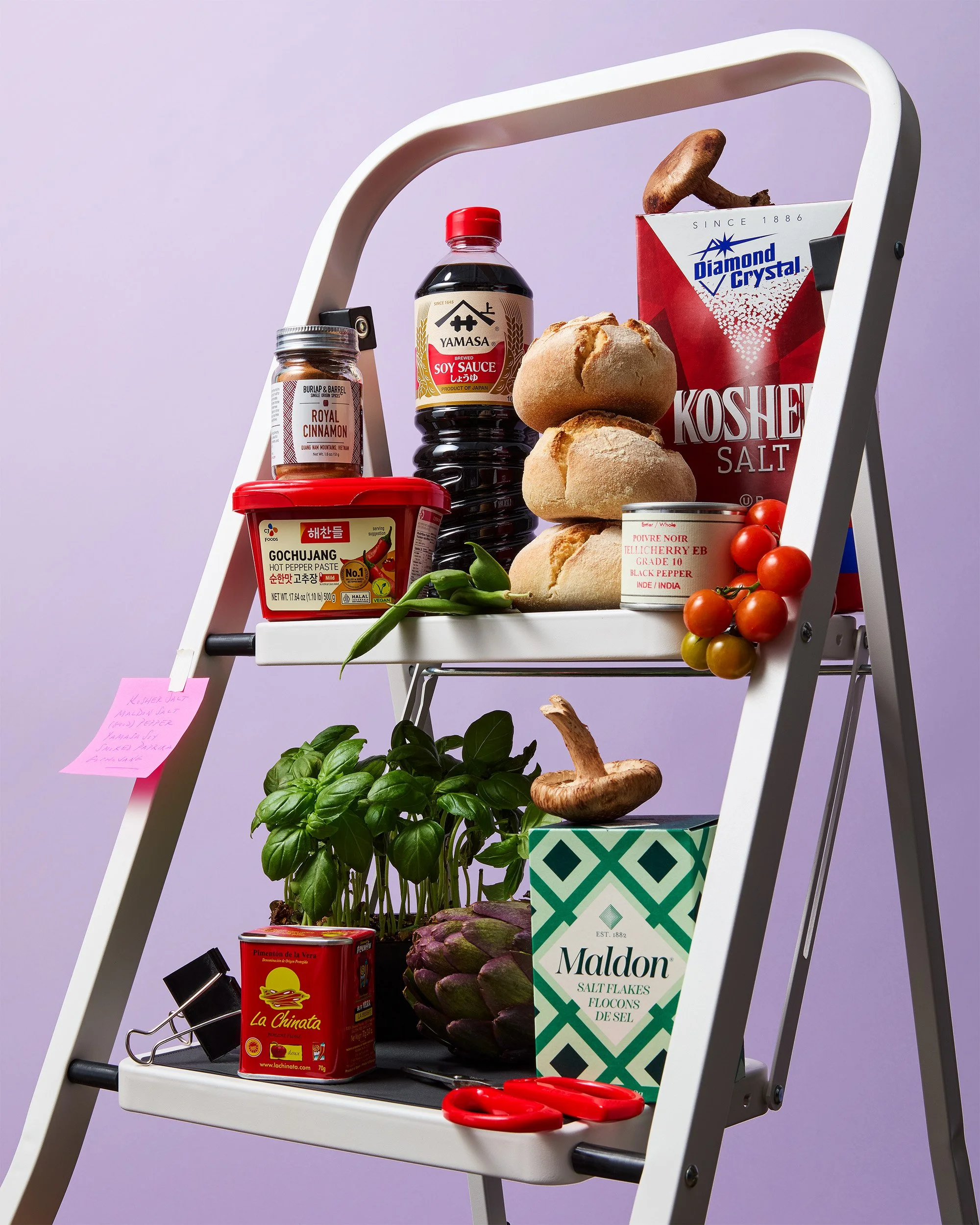 A two-tier white folding shelf holding various grocery items, including fresh herbs, artichokes, mushrooms, cherry tomatoes, a container of kimchi, salt flakes, ginger root, a bottle of soy sauce, a jar of cinnamon, a container of Gochujang spicy pas