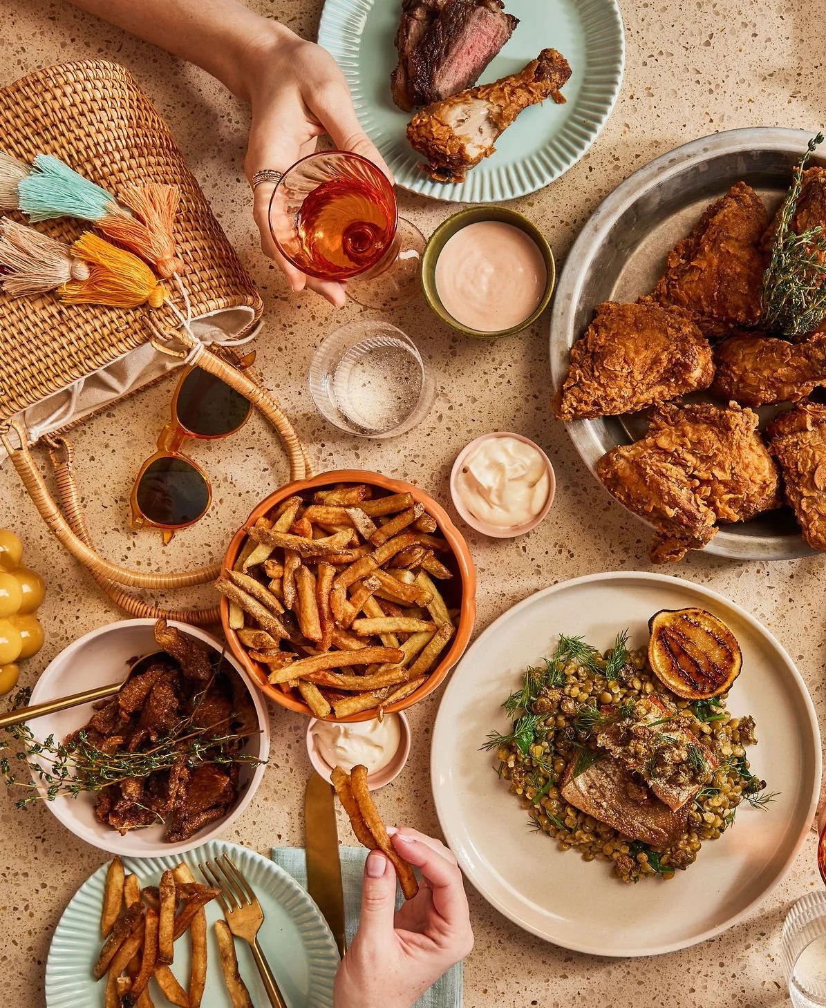 HEYDAYS RESTAURANT COOKBOOK at The June Motel. A styled photographed table spread with various dishes including fried chicken, steak, French fries, a salad with fish, and dipping sauces, along with a glass of iced tea and sunglasses.