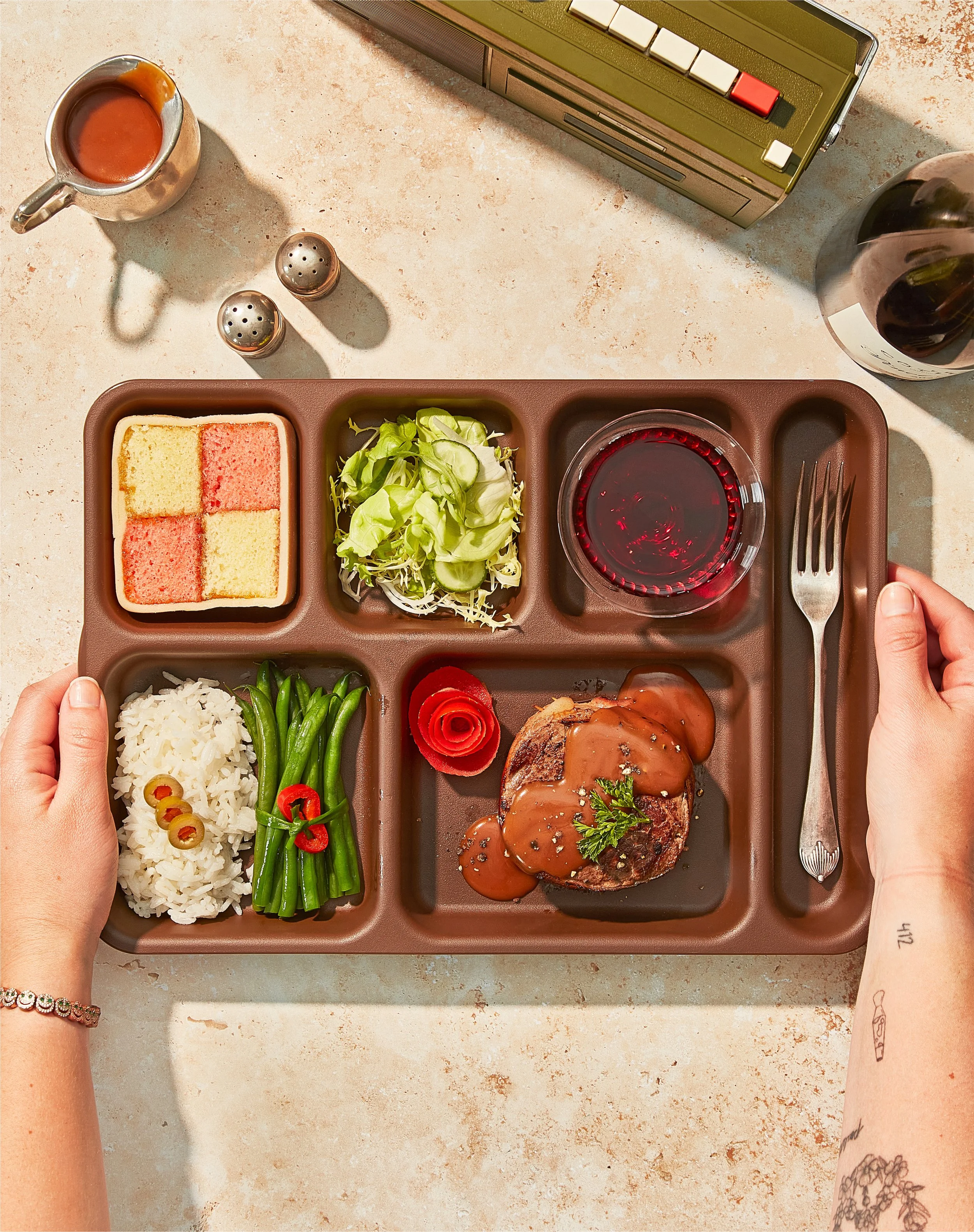 Tabletop creative photography prop styled and art directed by rayna marlee. A brown divided tray with a meal including rice, green beans, a grilled meat patty with sauce, a salad, a small plate of layered cake, a glass of red wine.