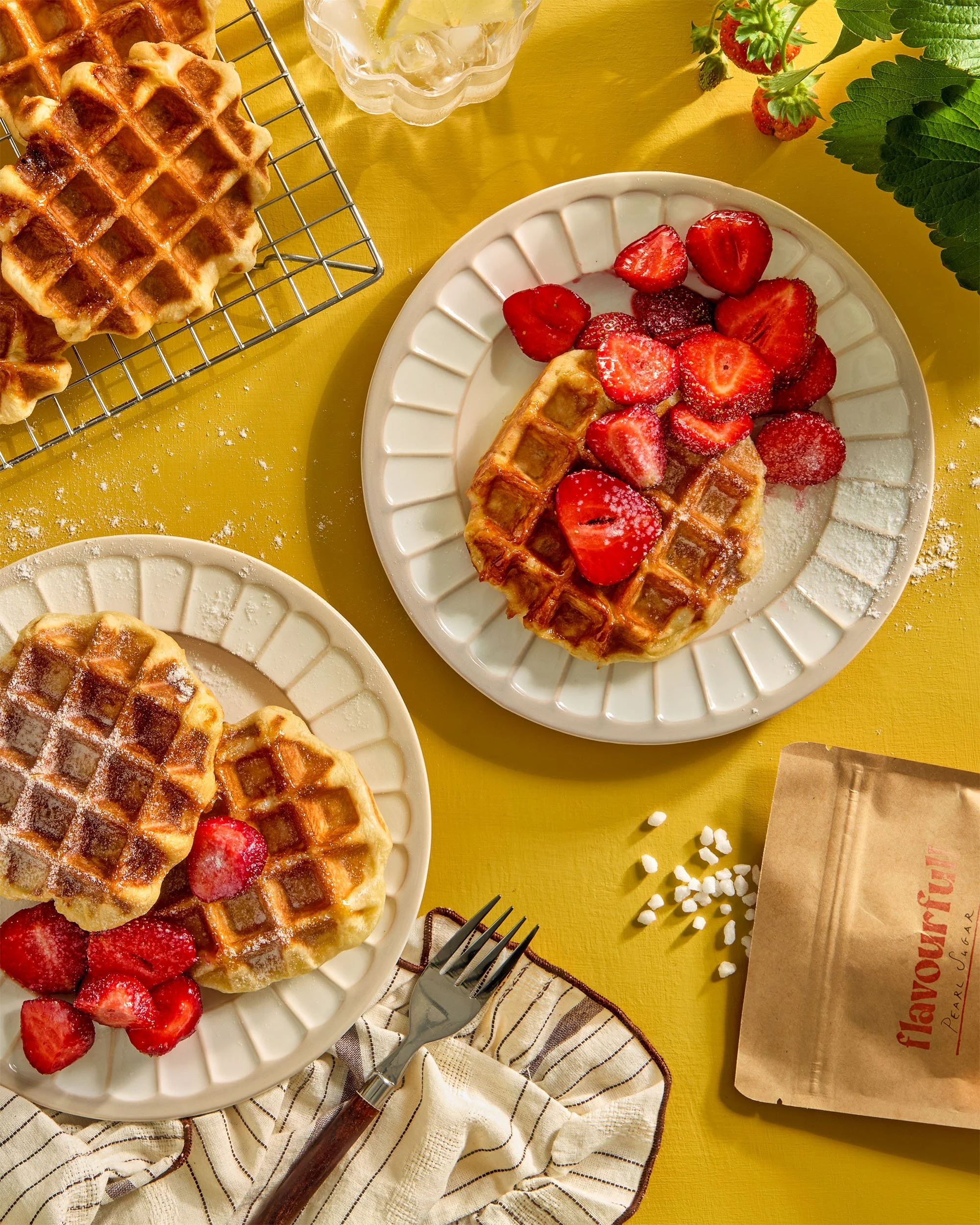 Tabletop creative photography prop styled by rayna marlee. Waffles topped strawberries, powdered sugar, and syrup on white plates, set on a yellow table with a knife, a fork, a glass of water with lemon, a paper bag, and some fresh strawberries.