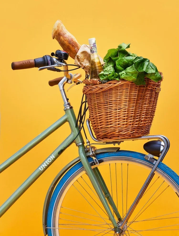 Still life creative photography prop styled and art directed by rayna marlee for Go Dutch Bikes. A green bicycle with a front basket filled with leafy greens, a baguette, a bottle, and a paper bag, set against a yellow background.