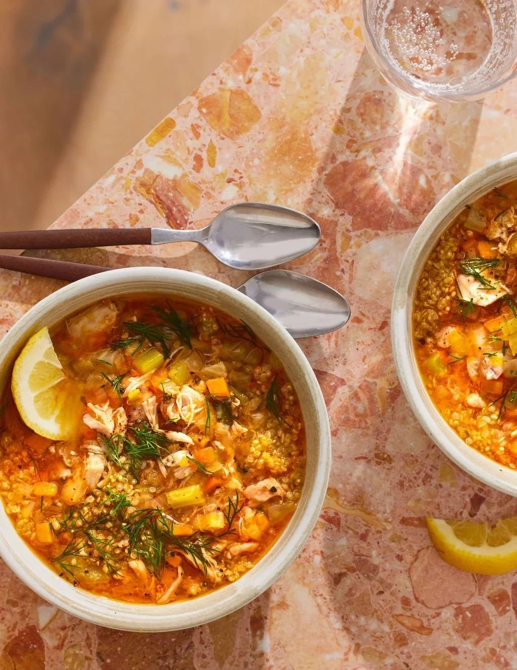 Cover of a cookbook titled "The Young Forever Cookbook" by Mark Hyman, MD, art directed and prop styled by rayna marlee bowls of chicken and vegetable soup garnished with lemon wedges and herbs, on a pink marble table with spoons and a glass of water