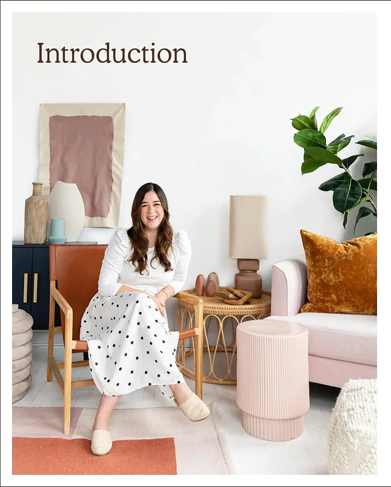 A woman Home Decor Youtuber Alexandra Gater Book sitting in a modern living room with a white wall that has the word 'Introduction' written on it. She is smiling and wearing a white top and a white skirt with black polka dots. 