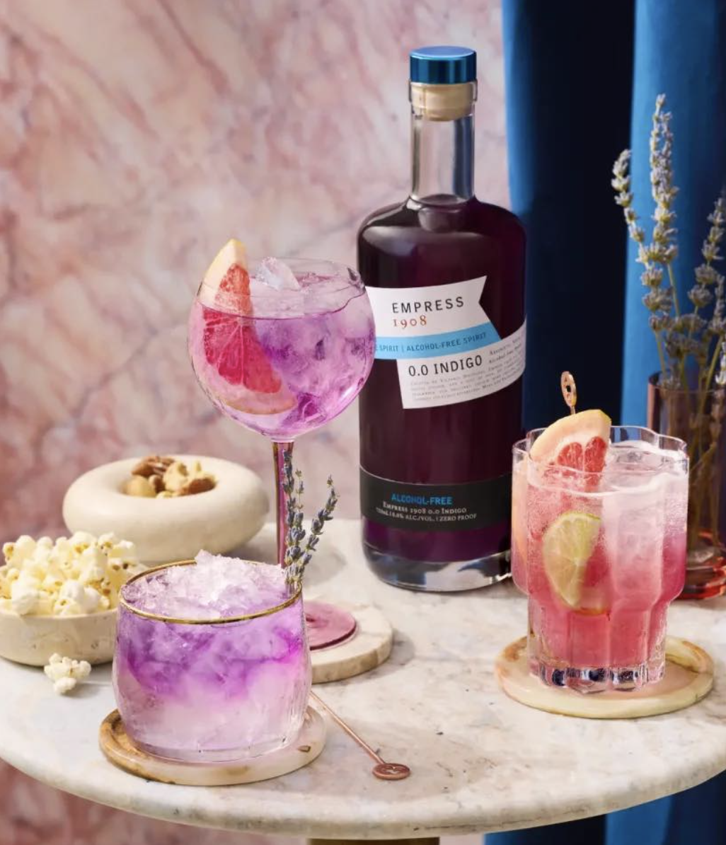 A variety of colorful cocktails on a marble table, with a bottle of alcohol-free spirit labeled 'Empress 1908', and bowls of popcorn and nuts, set against a pink and blue background with flowers.