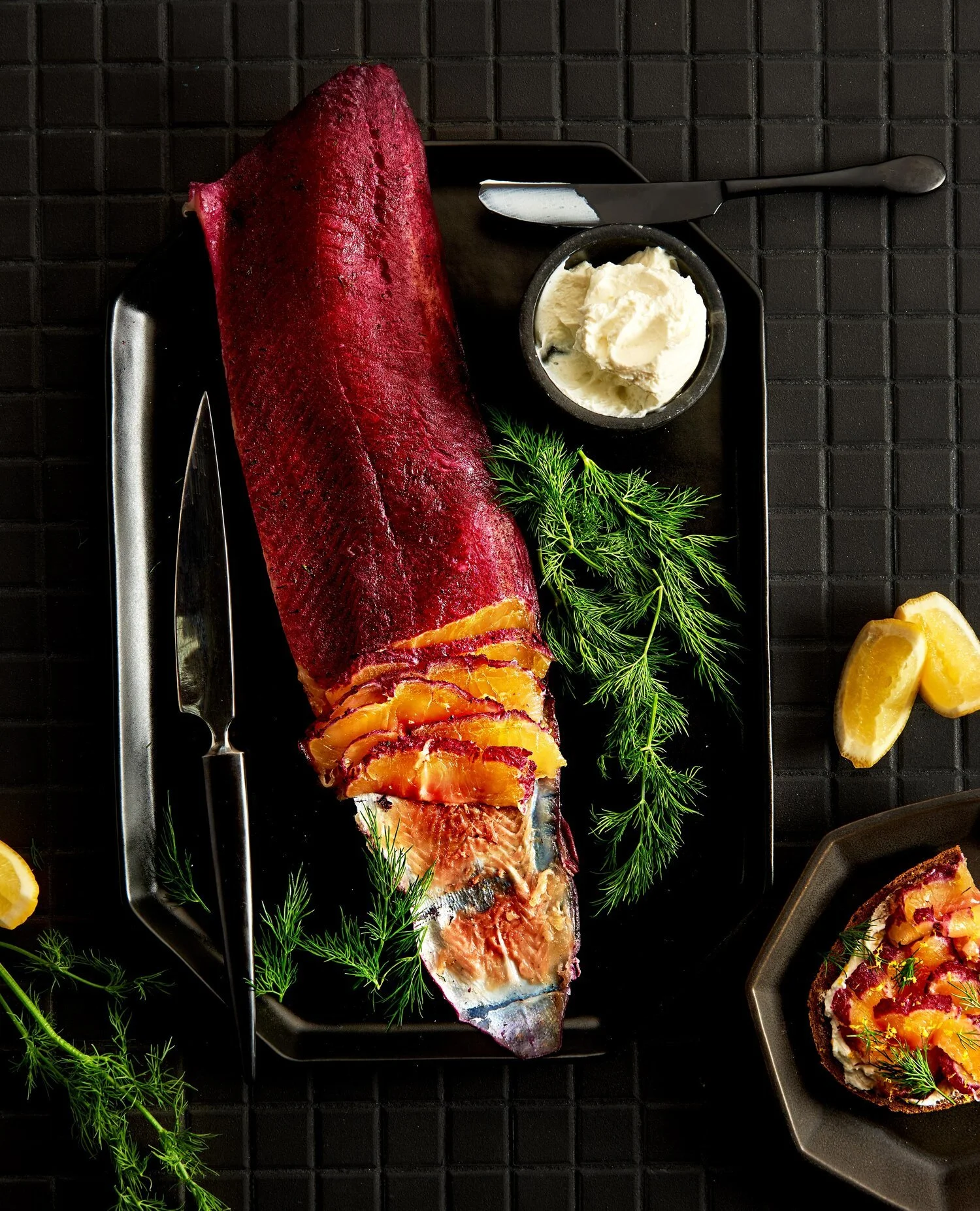 Food creative photography prop styled and art directed by rayna marlee for cookbook. A smoked fish fillet garnished with herbs, lemon lemon wedges, and a side of butter on a black tray.