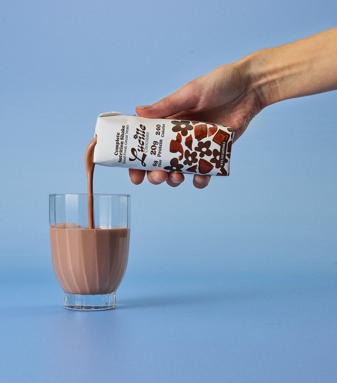 Still life creative photography prop styled by rayna marlee for Lucille. A hand pouring chocolate milk from a carton into a glass against a light blue background.