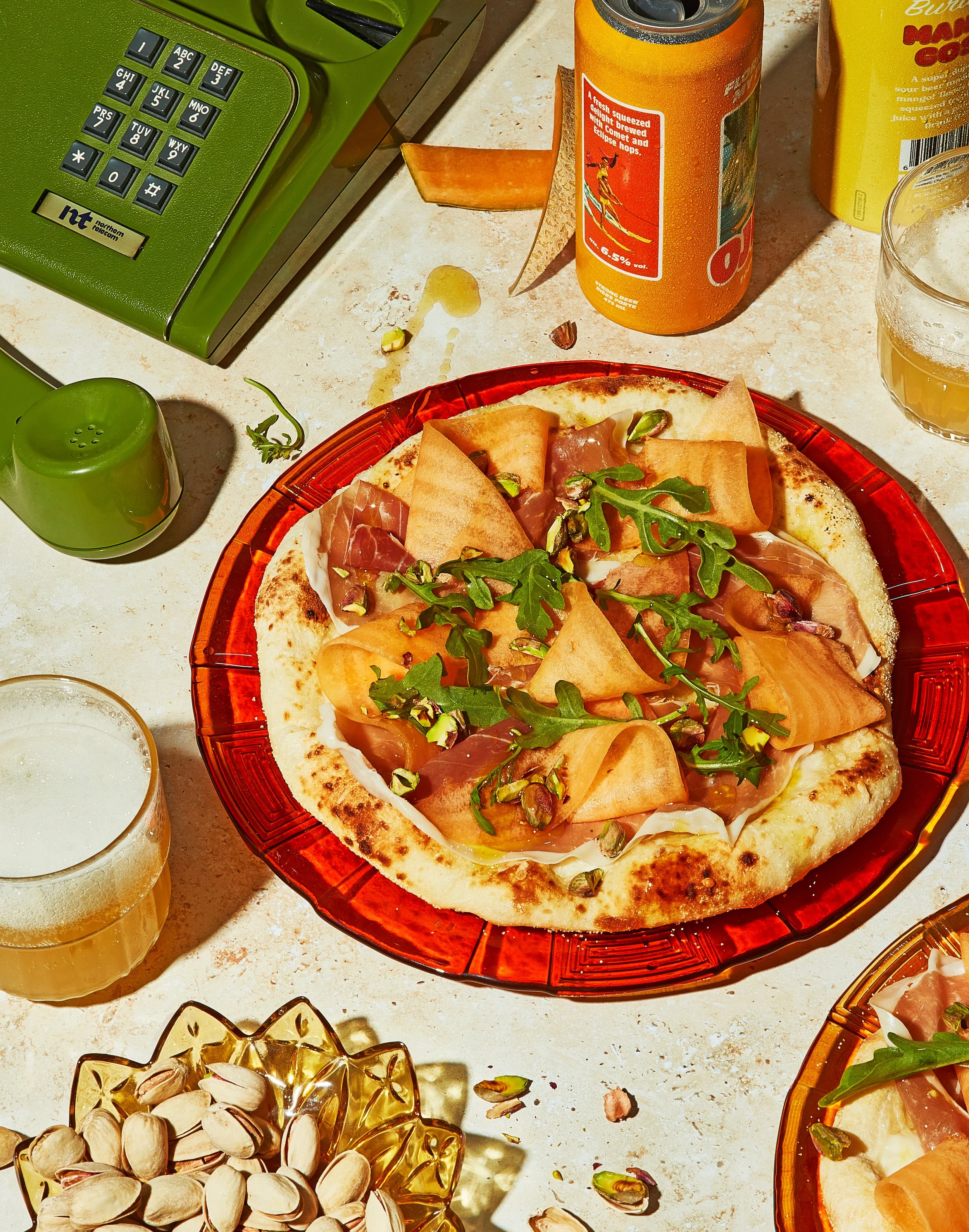 Food creative photography prop styled and art directed by rayna marlee. A slice of pizza with prosciutto, arugula, and pistachios on a red plate, surrounded by drinks, snacks, and a vintage green rotary phone on a light-colored table.