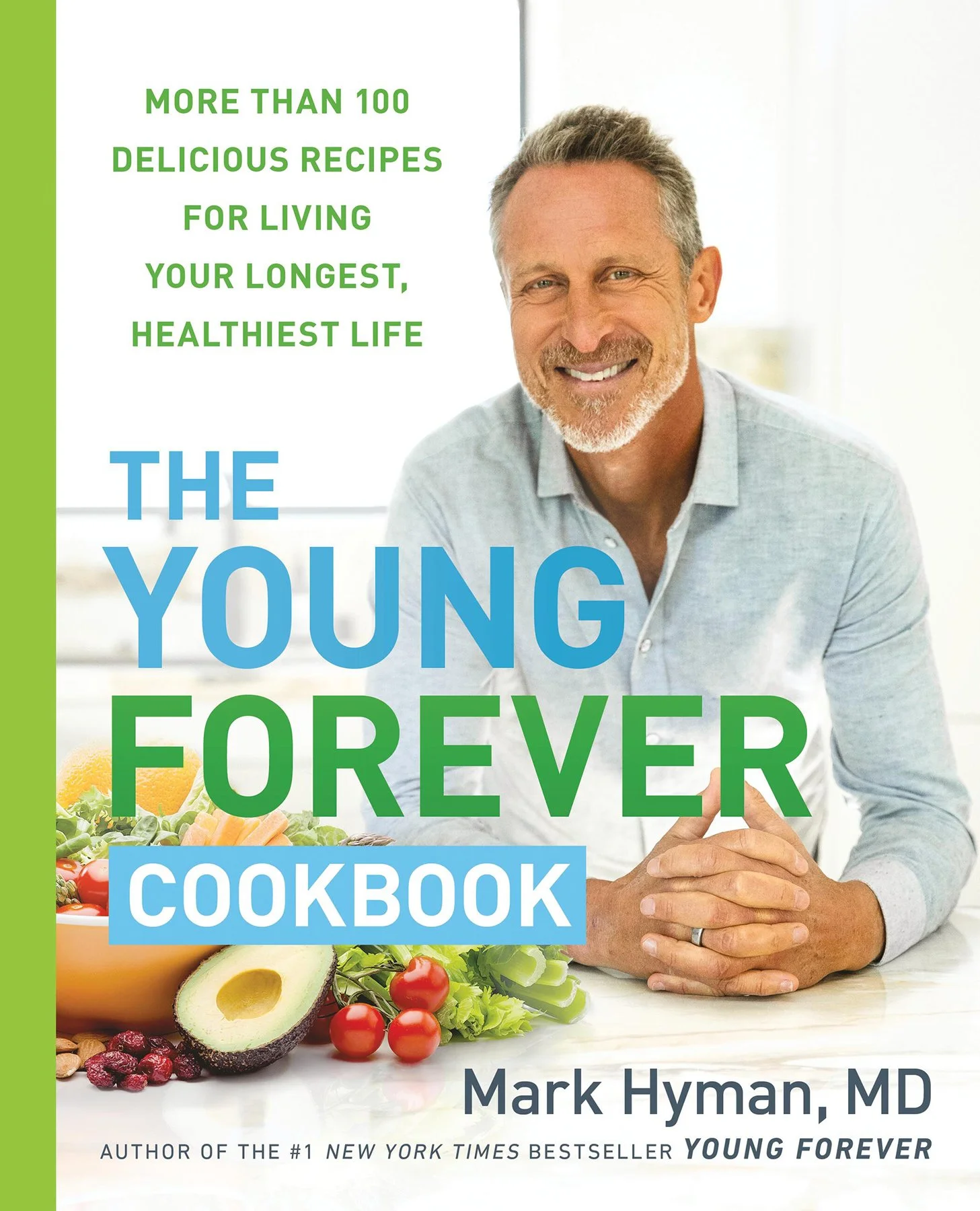 Cover of a cookbook titled "The Young Forever Cookbook" by Mark Hyman, MD, art directed and prop styled by rayna marlee. Mark sitting at a table with fresh vegetables and fruits like avocados, tomatoes, and lettuce in the foreground.