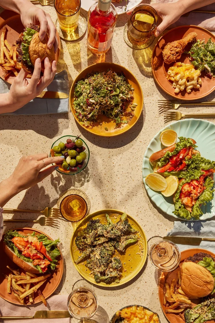 Food creative photography prop styled and art directed by rayna marlee for Hey Days Coobook. A variety of dishes including burgers, fries, salads, fried chicken, broccoli, a bowl of mixed grapes, and drinks on a table during a meal.