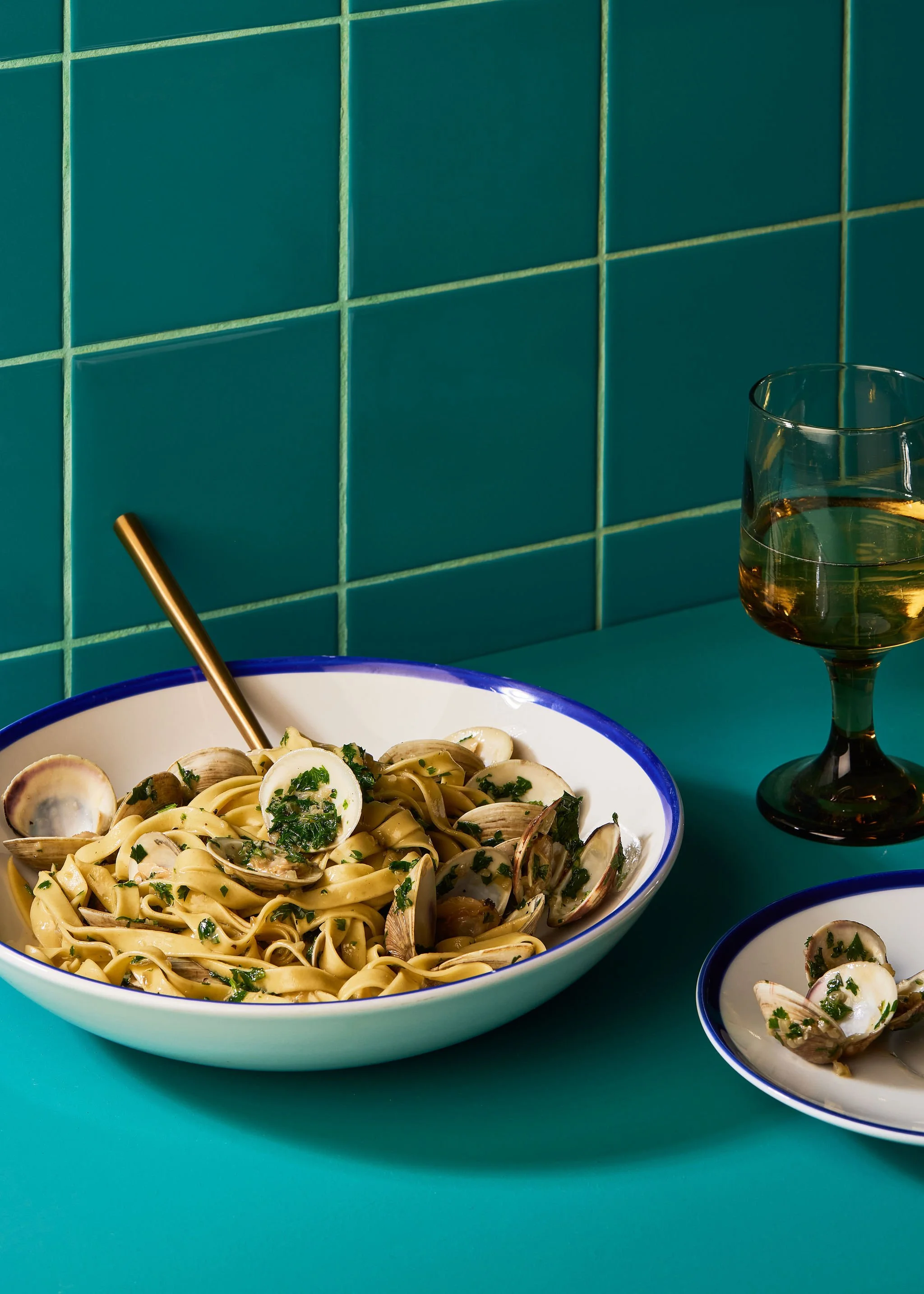 Still life creative photography prop styled and art directed by rayna marlee. Hey Days Cookbook. A bowl of pasta with clams and chopped herbs, a glass of white wine, and a smaller plate with clams on a teal table against teal tiled wall.