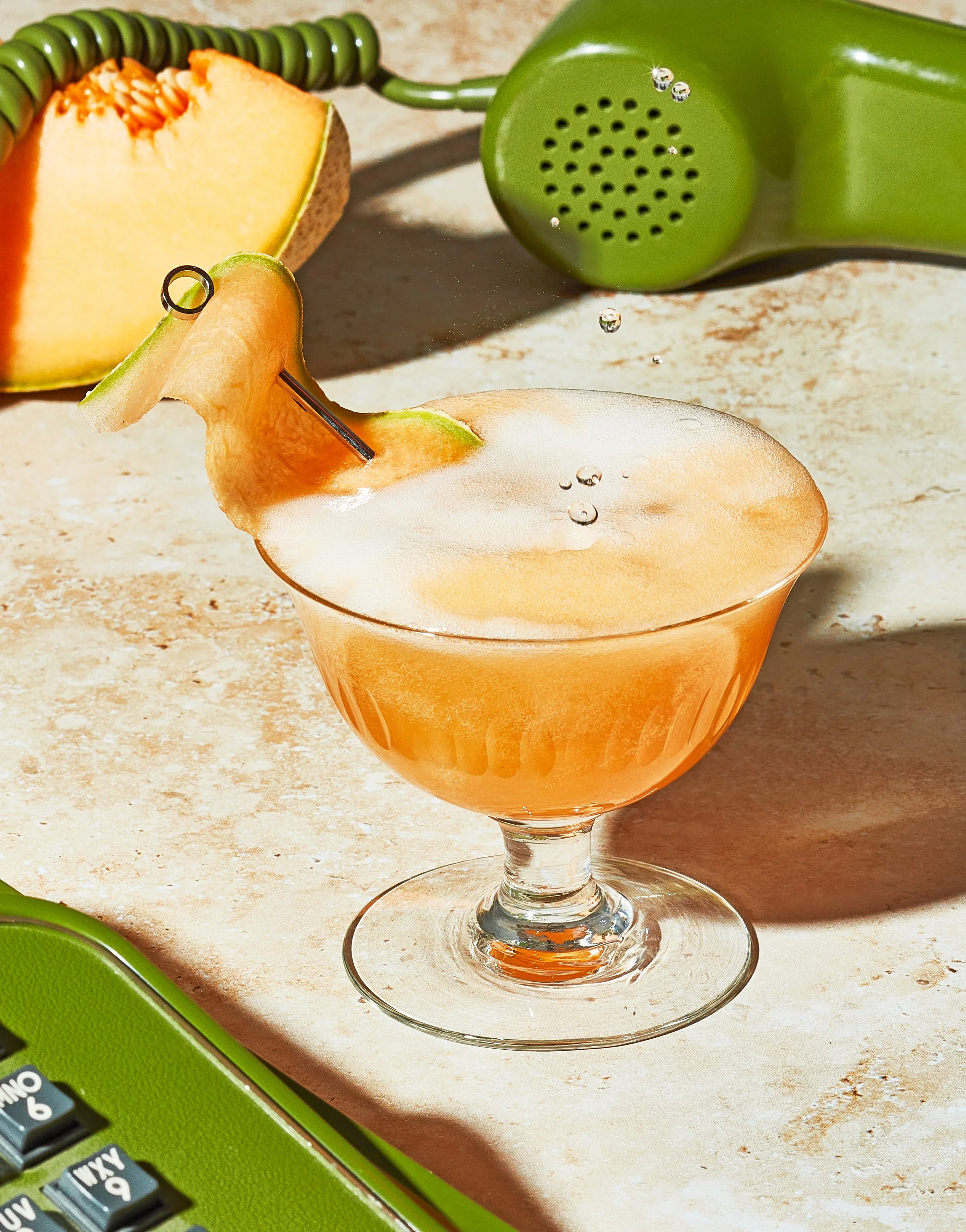  Still life creative photography prop styled and art directed by rayna marlee. A peach-flavored cocktail garnished with a slice of peach on the rim, served in a vintage glass with a straw. The background has a sliced cantaloupe, a green rotary phone.