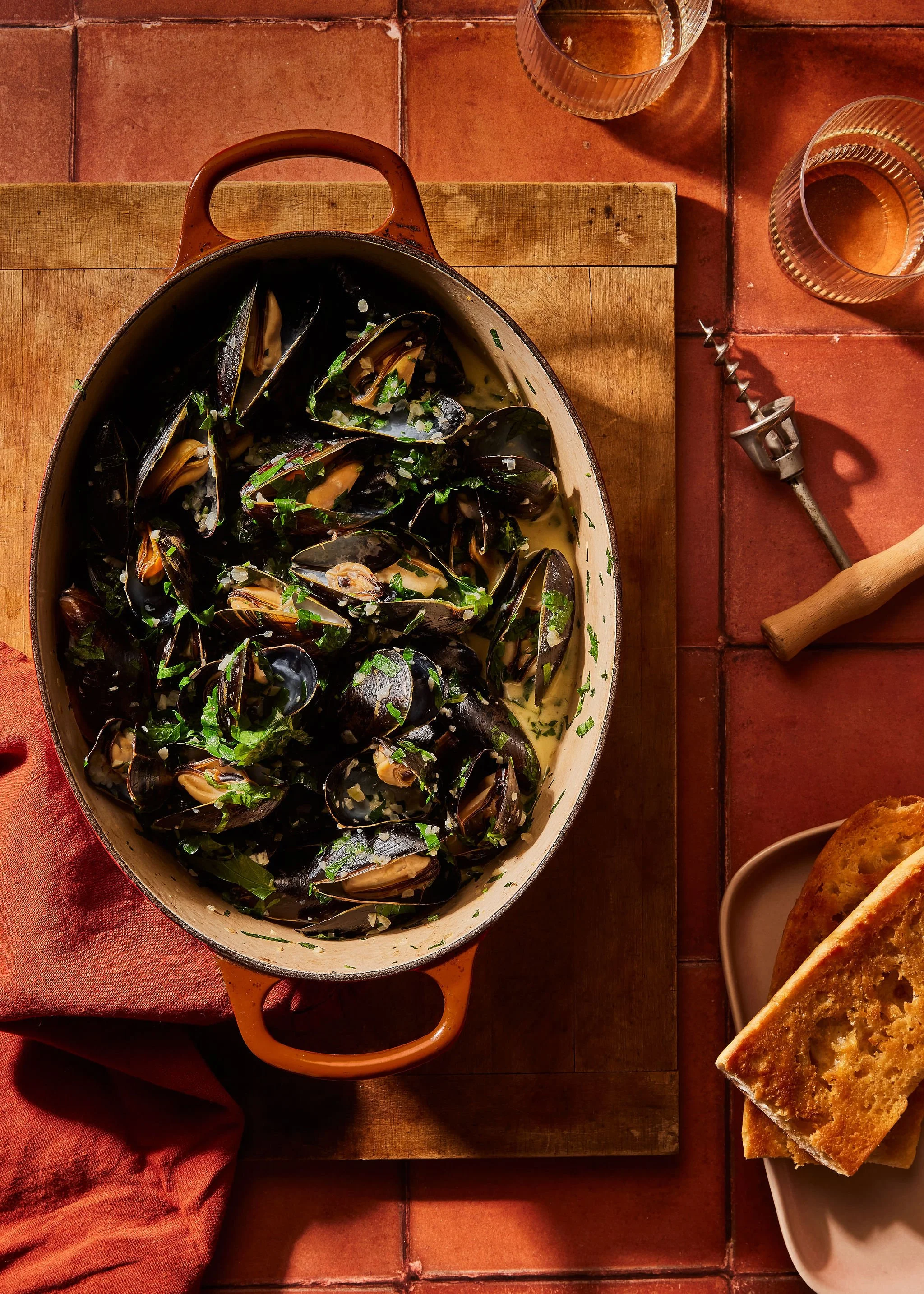 A casserole dish filled with cooked mussels in a creamy sauce, garnished with chopped herbs, placed on a wooden board. To the right, there are two glasses of white wine, a corkscrew, a wooden-handled tool, and a sliced loaf of bread on a beige plate.
