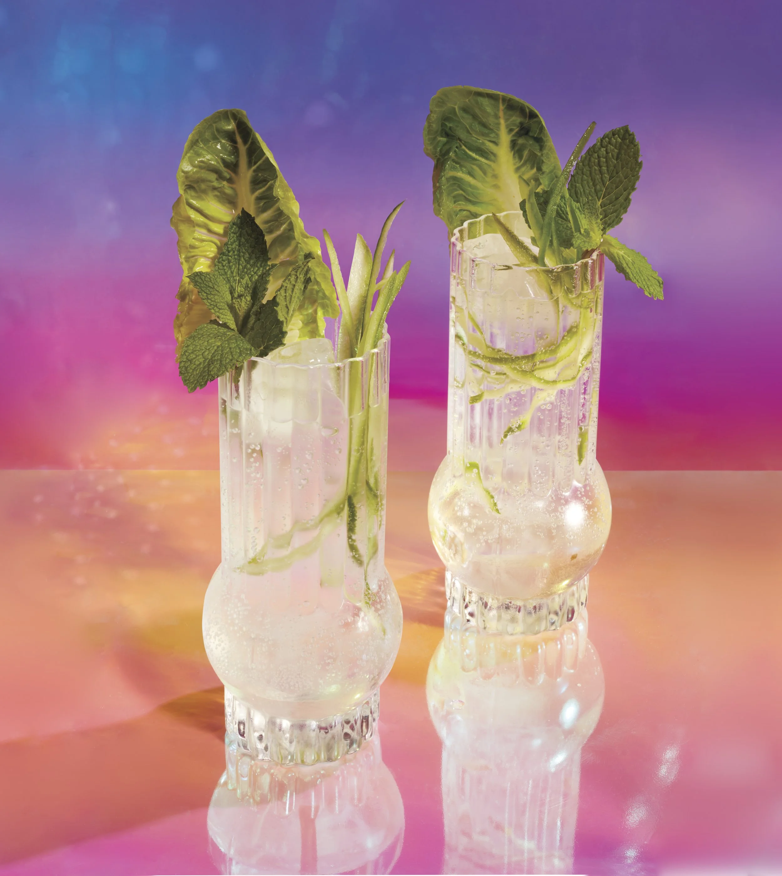 Drink photography prop styled by rayna marlee for Chatelaine Magazine. Two glasses with clear beverage, garnished with mint leaves, lettuce, and cucumber slices, set against a colorful, sunset-inspired background.