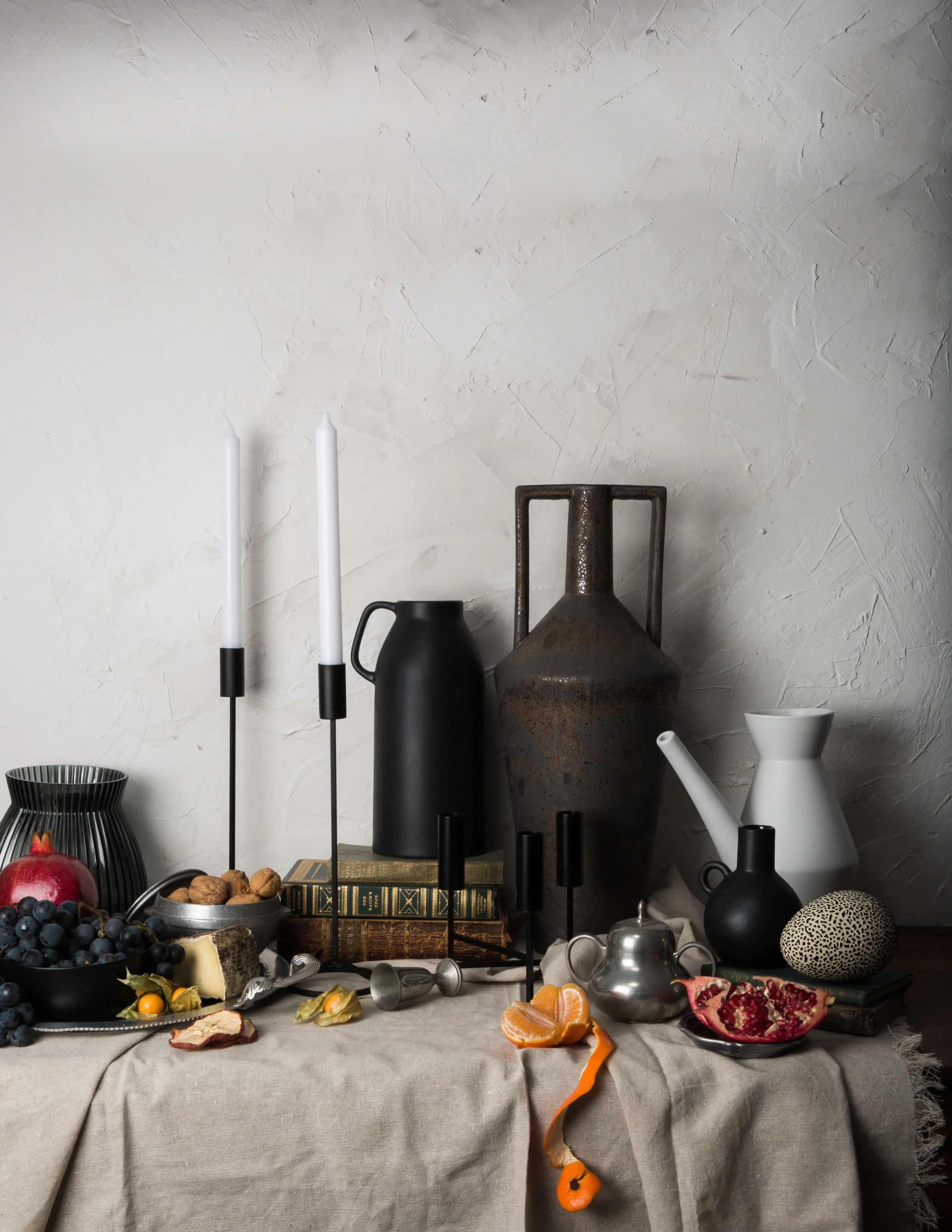 Still life creative photography prop styled and art directed by rayna marlee. Still life arrangement of various objects on a draped table, including fruits, vases, candlesticks, books, and decorative items, against a textured white wall background.