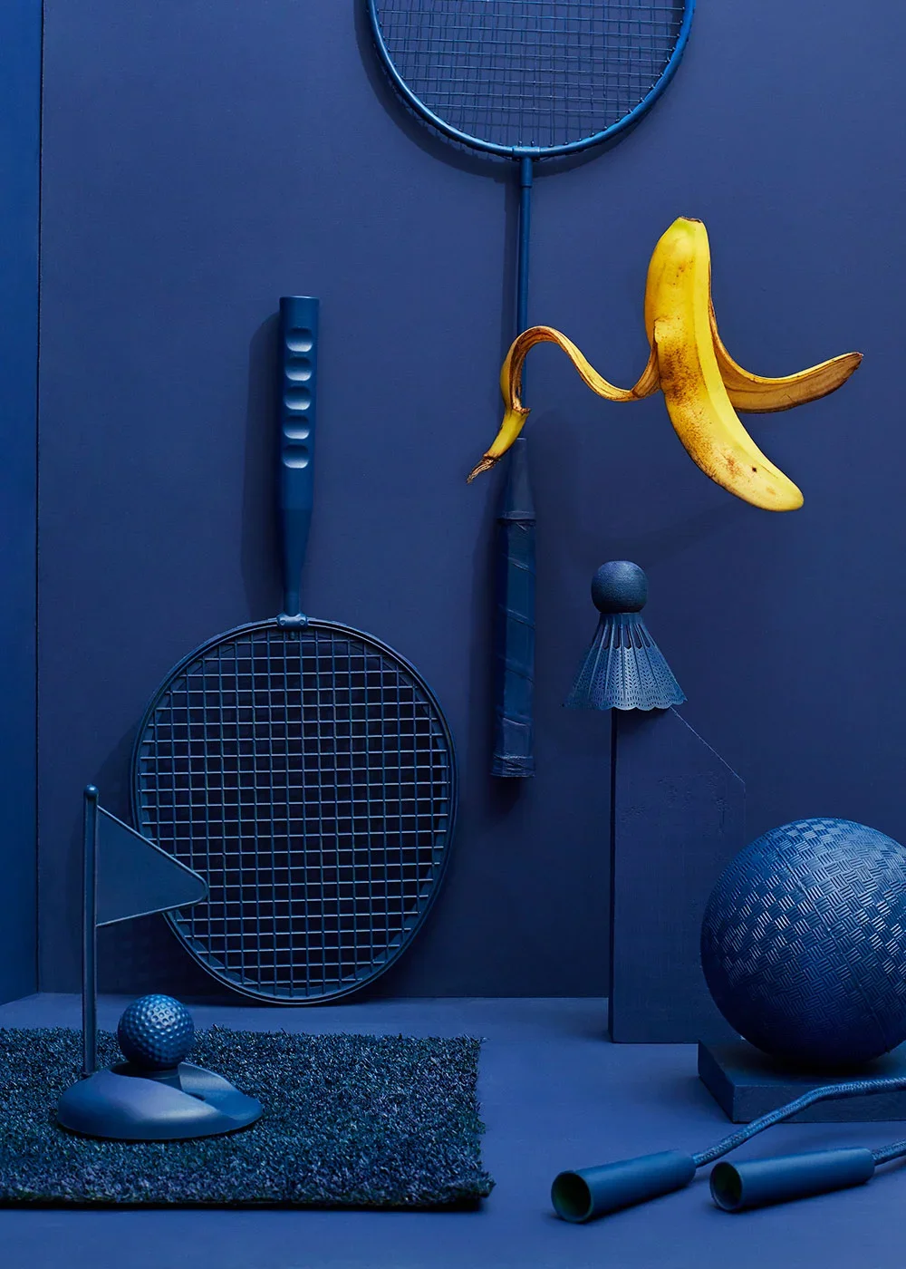 Still life creative photography prop styled and art directed by rayna marlee. Sports equipment and a banana hanging on a dark blue wall, including tennis rackets, a shuttlecock, a golf ball on a tee, a large textured ball, and two batons.