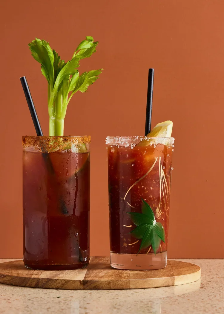 Drink creative photography prop styled and art directed by rayna marlee for Heydays Magazine. Two tall glasses of Bloody Mary cocktails on a wooden tray, one garnished with celery and the other with a lemon wedge, against an orange background.