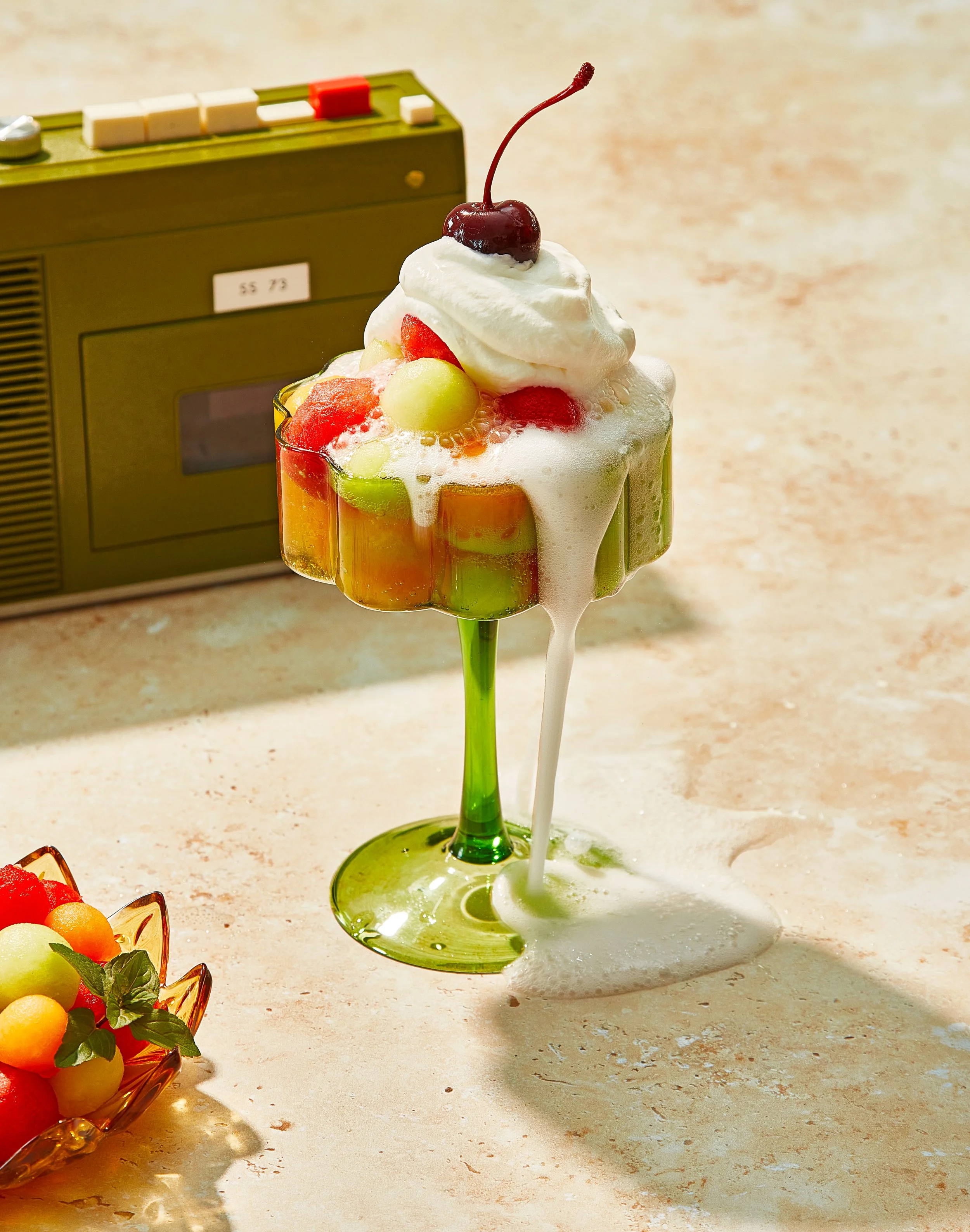 Drink creative photography prop styled and art directed by rayna marlee. A tall, green cocktail glass filled with colorful fruit chunks and topped with whipped cream, a cherry, and fruit slices, with foam overflowing onto the surface.