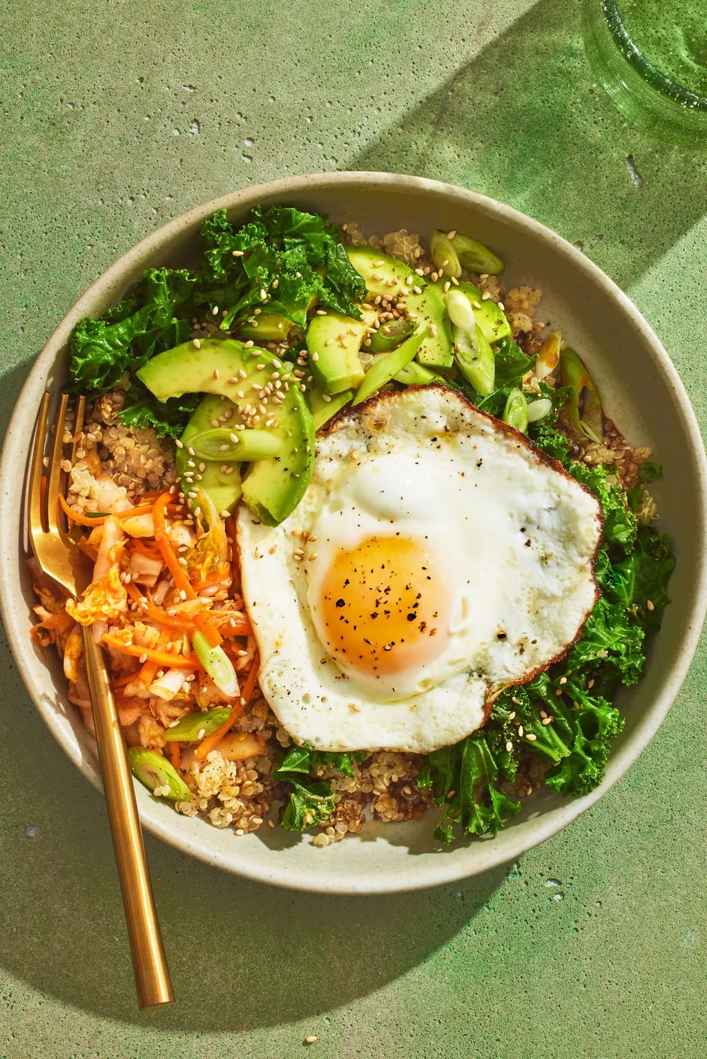 Cover of a cookbook titled "The Young Forever Cookbook" by Mark Hyman, MD, art directed and prop styled by rayna marlee. A bowl of quinoa salad topped with avocado slices, kale, and a fried egg, with a gold fork on the side and a glass
