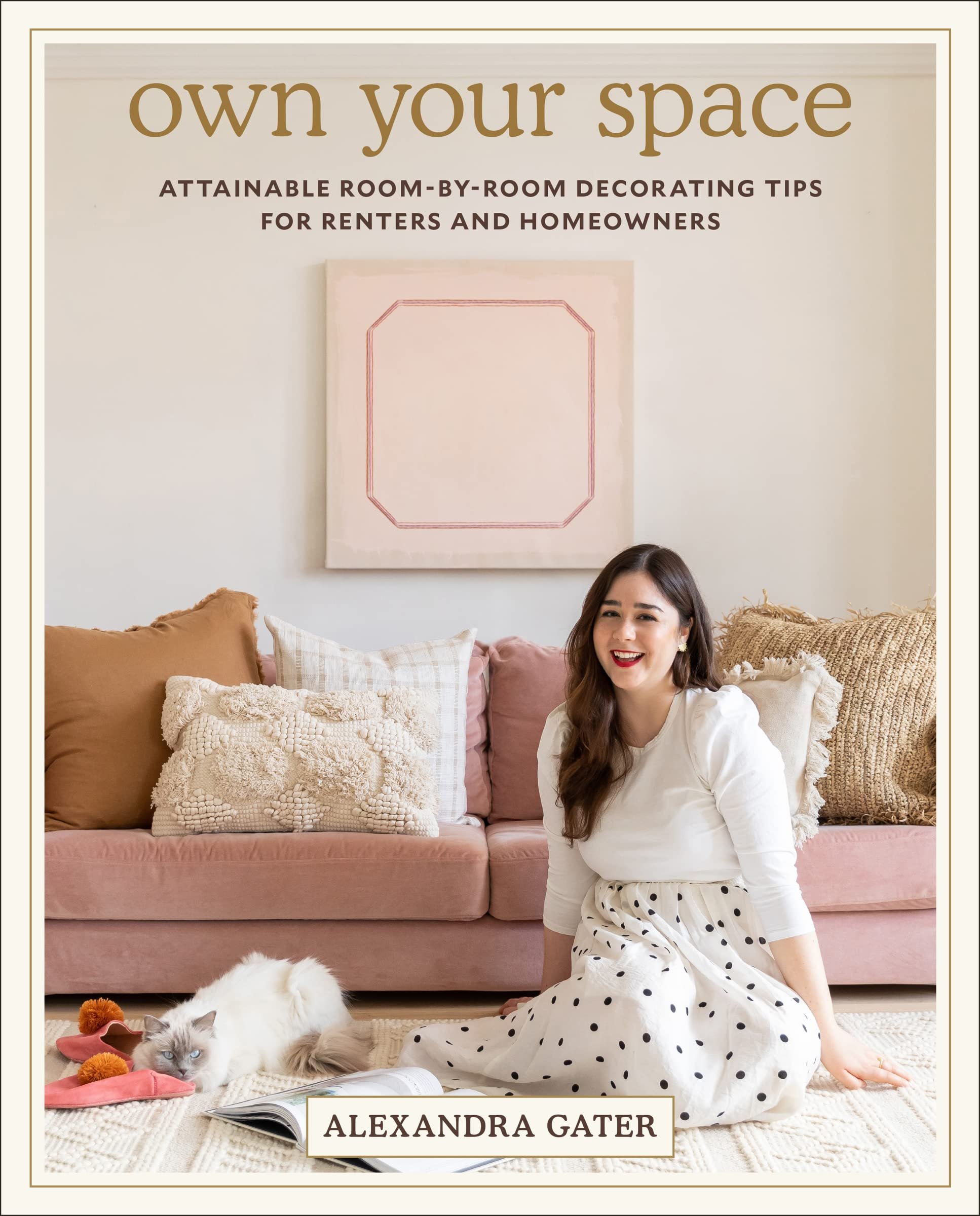 A woman sitting on the floor in a living room with a sofa and decorative pillows, a cat lying nearby, and a book in front of her. The cover of a book or magazine has the author’s name, Alexandra Gater, on it. There is wall art behind her and a framed